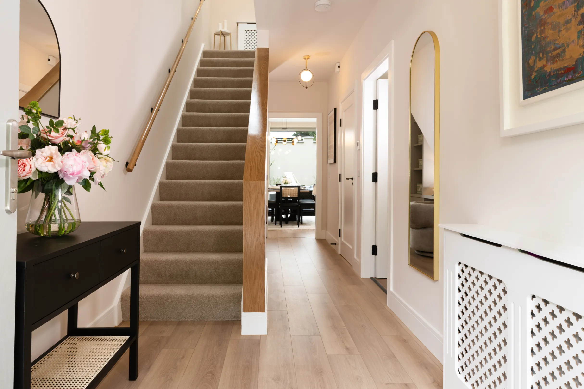 A bright, modern hallway features a staircase, dark console table with flowers, a long mirror, and a peek into a dining area.
