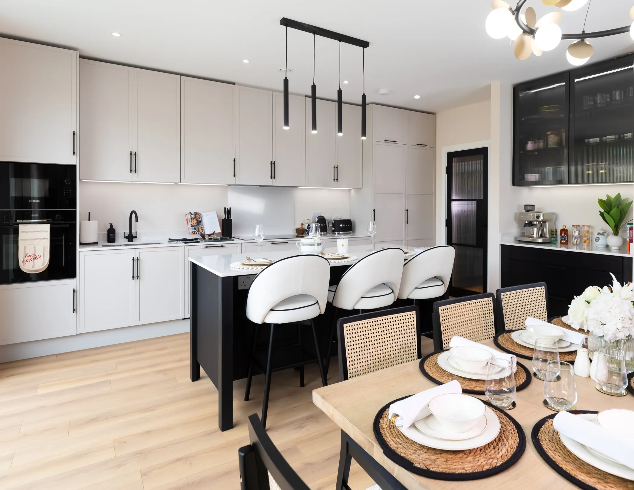 A modern kitchen with a black island and light wood cabinets, a dining table set for a meal, and decorative lighting.
