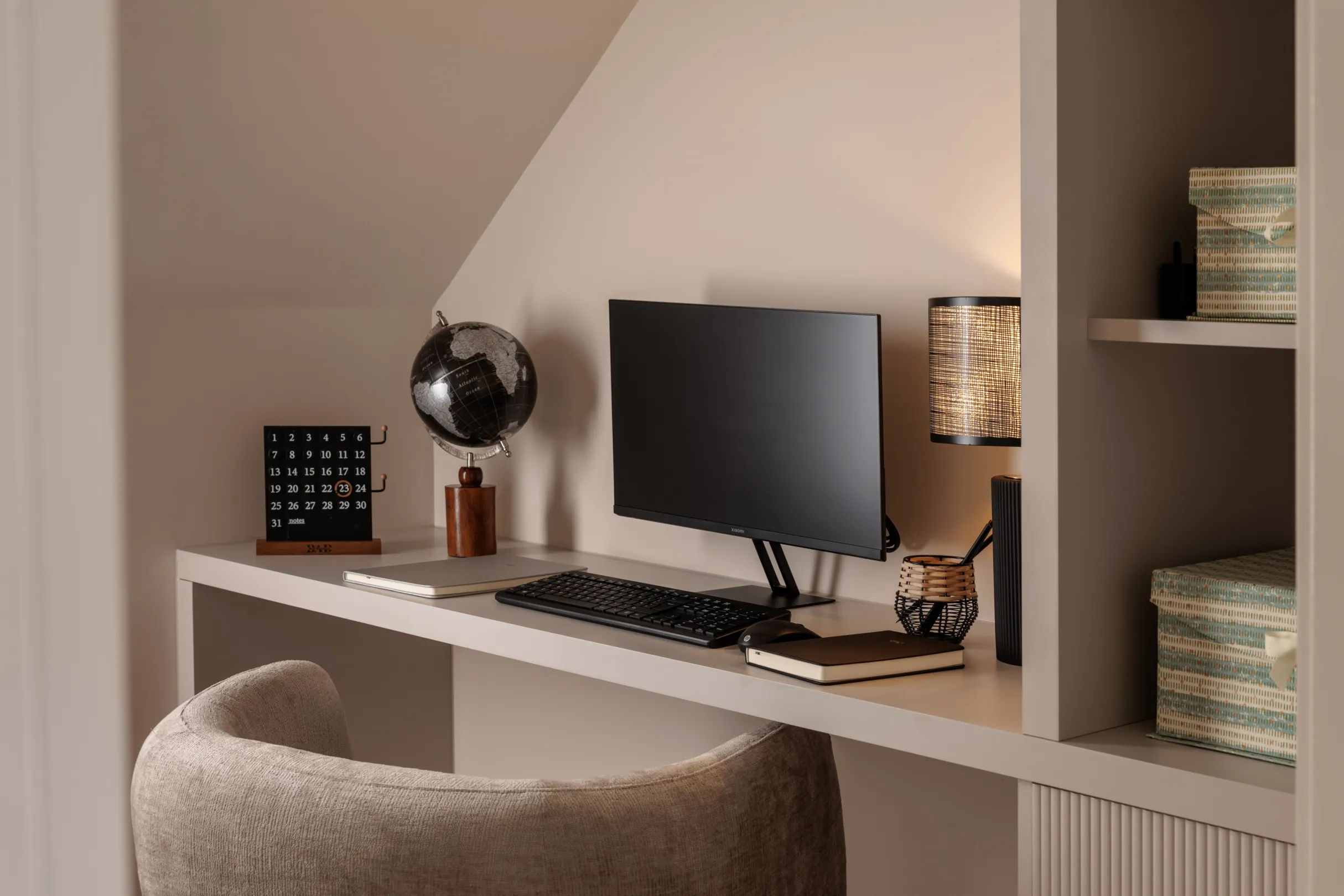 A neutral-toned home office setup features a desk with a computer, globe, calendar, and a comfortable chair.
