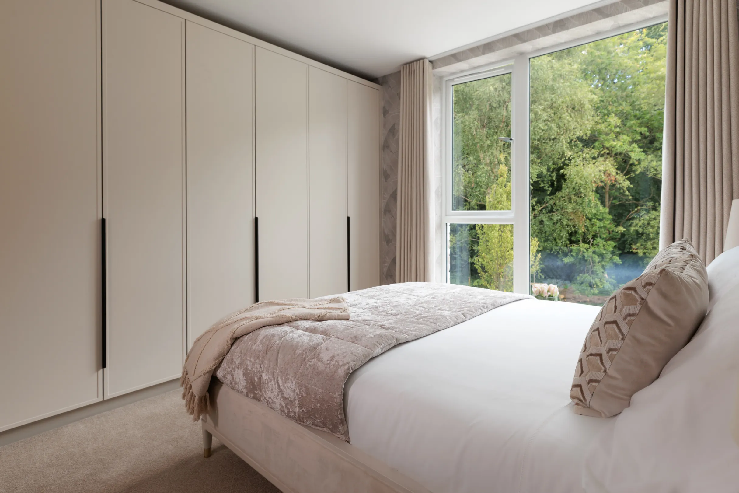 A bedroom features a large bed with a decorative pillow and blanket, positioned near a bright window with a view of lush greenery, next to a light-colored wardrobes.