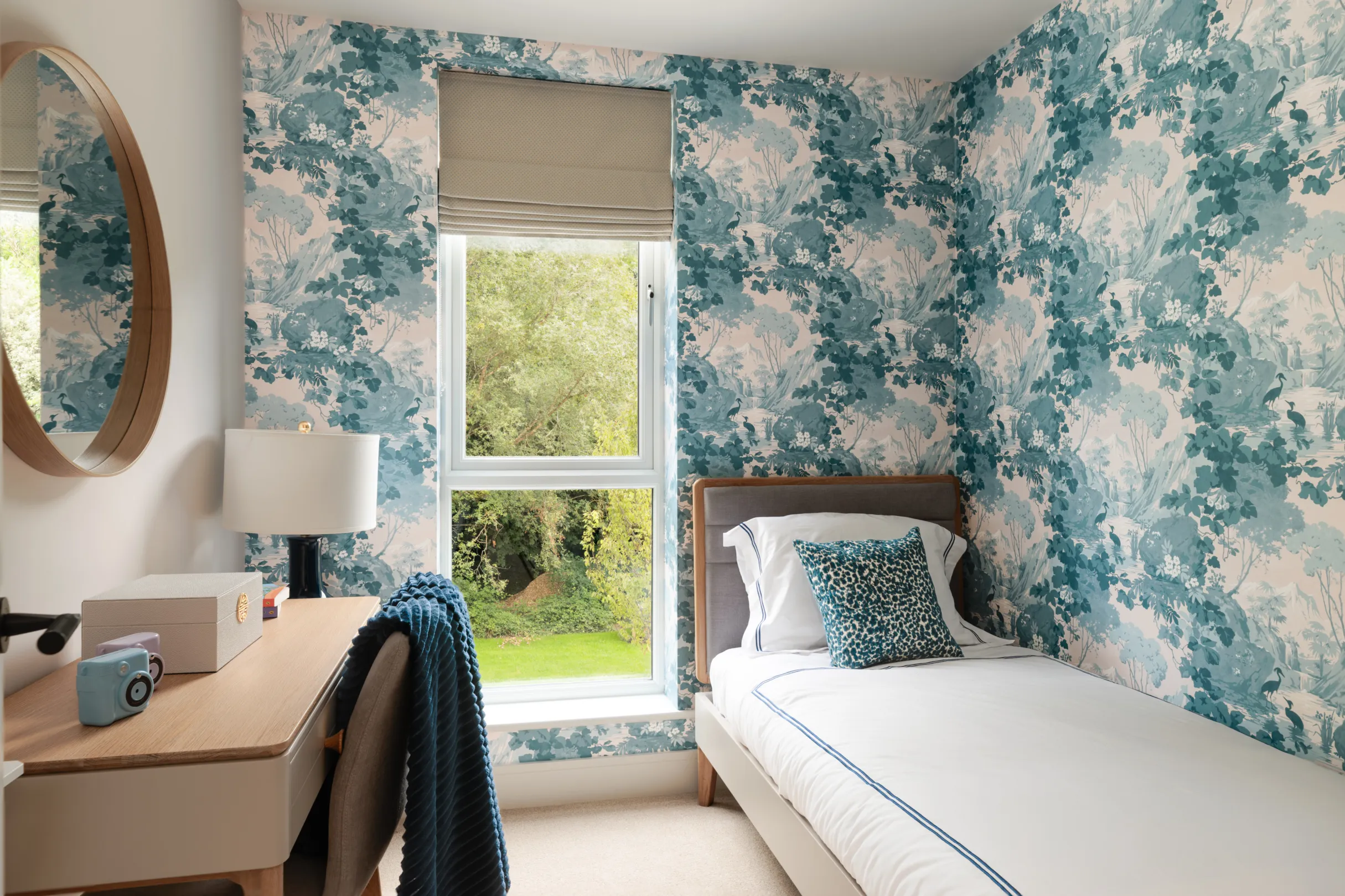 A bedroom featuring a desk, a single bed with patterned throw pillow, a round mirror, and blue and white patterned wallpaper.