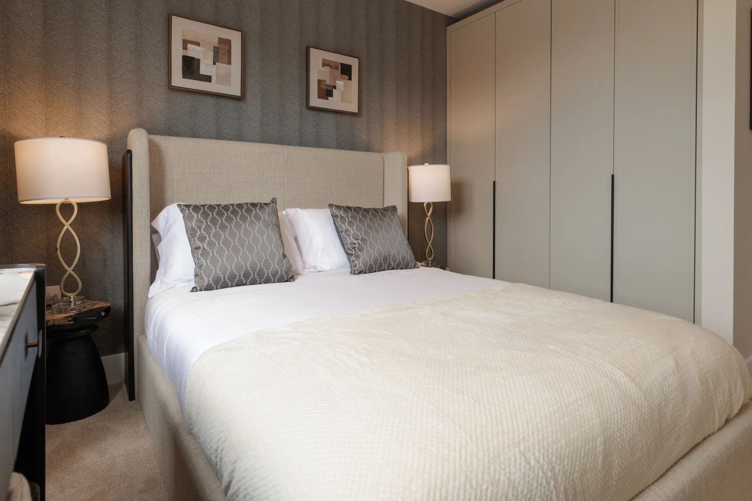 A bedroom features a bed with decorative pillows, lamps on bedside tables, and a wardrobe in the background.