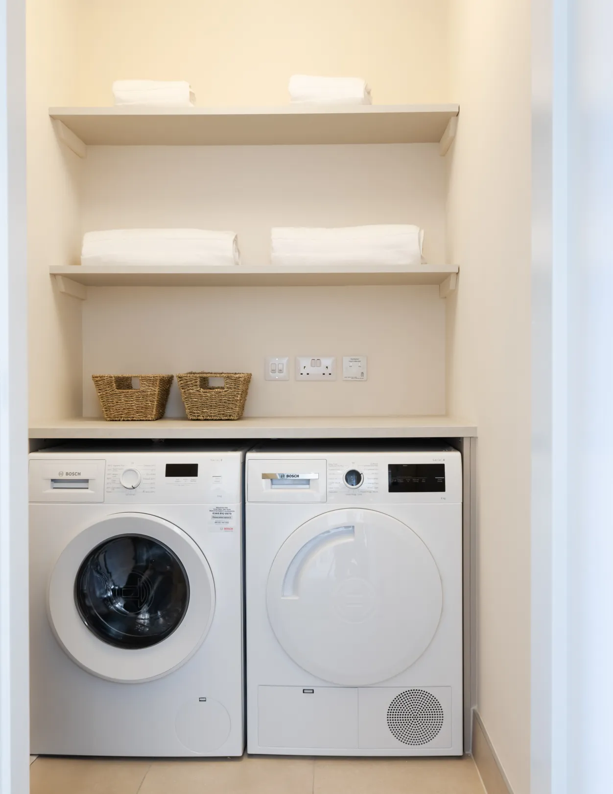 A laundry room features a white washing machine and dryer under shelves with neatly folded towels and woven baskets.