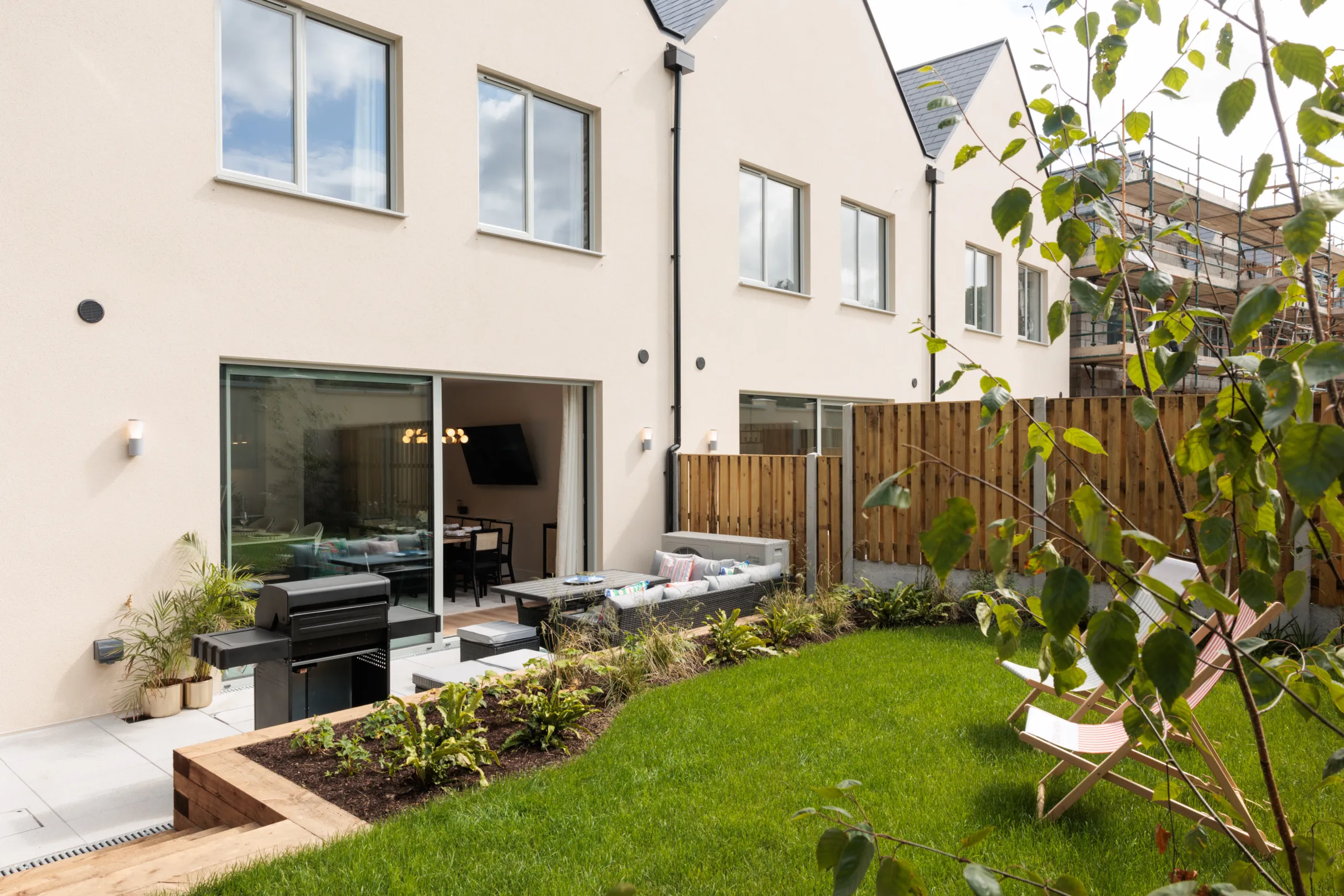 An outdoor patio setup features a wicker table and sectional sofa with colorful cushions, a grill, and lounge chairs against a garden backdrop.