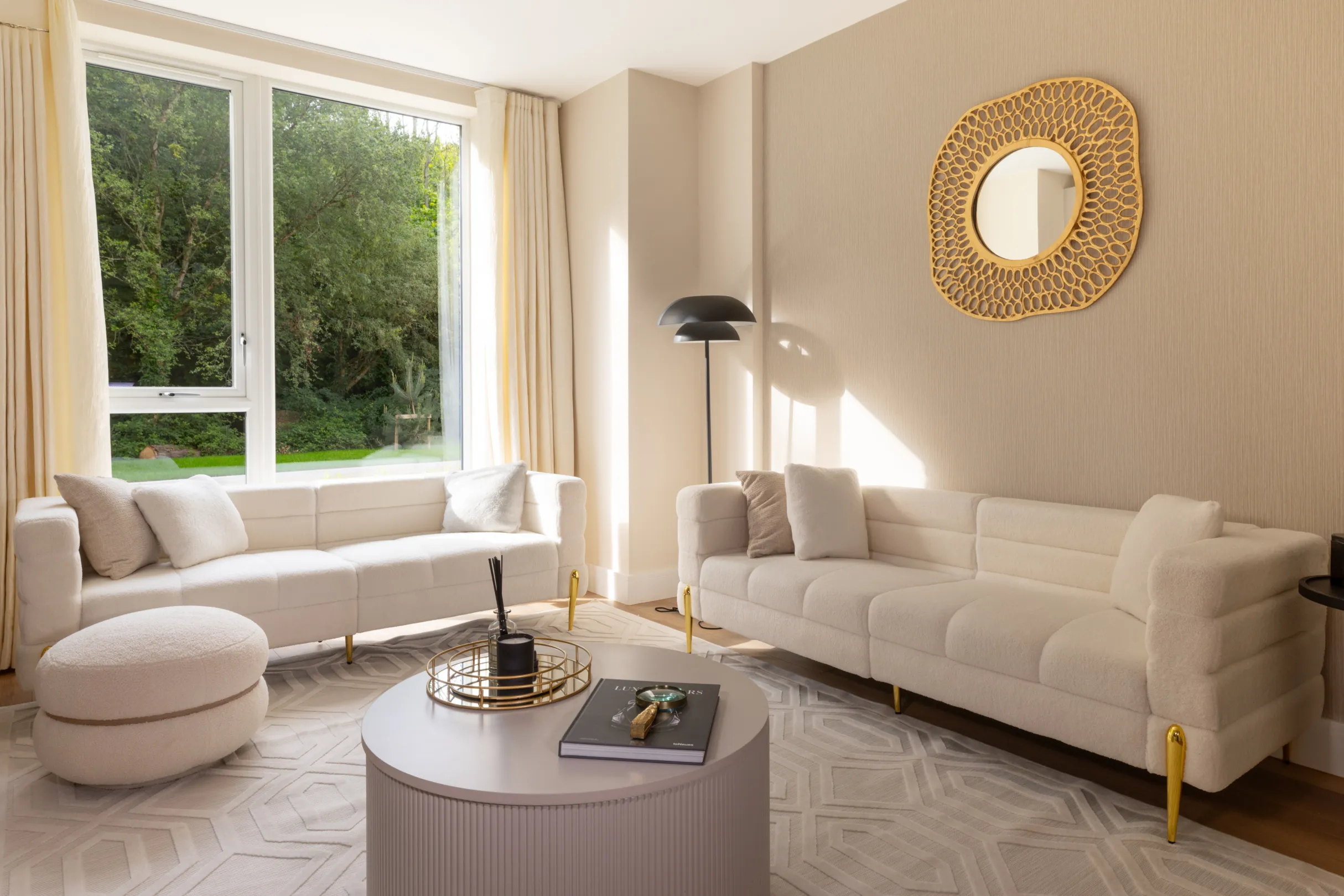 A bright living room features two cream-colored sofas, a round coffee table, an ottoman, and a decorative mirror.