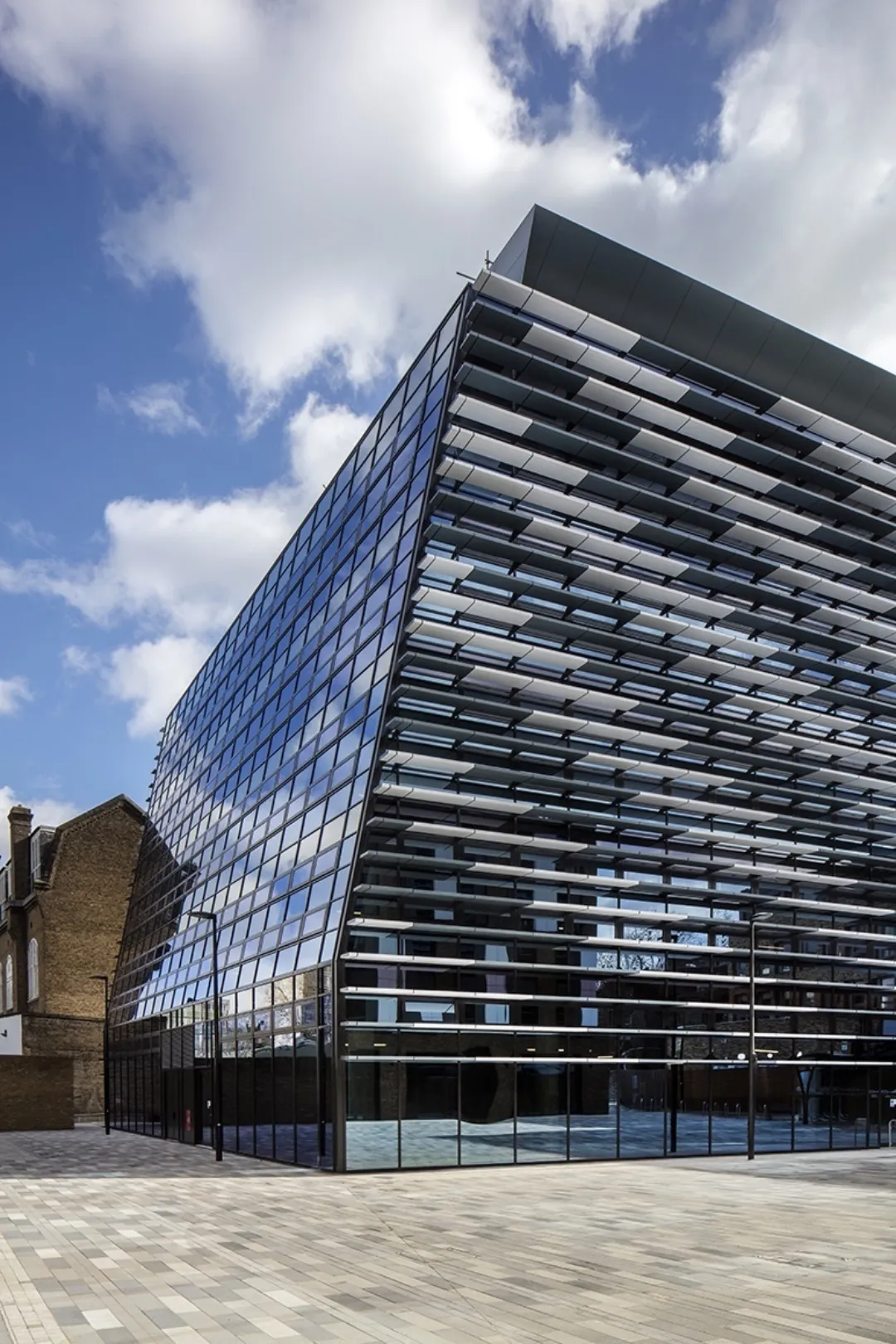 Striking facade of a modern building glazed on one side with solar shading on the other, an old brick construction home is in the background, with brick paving in the foreground