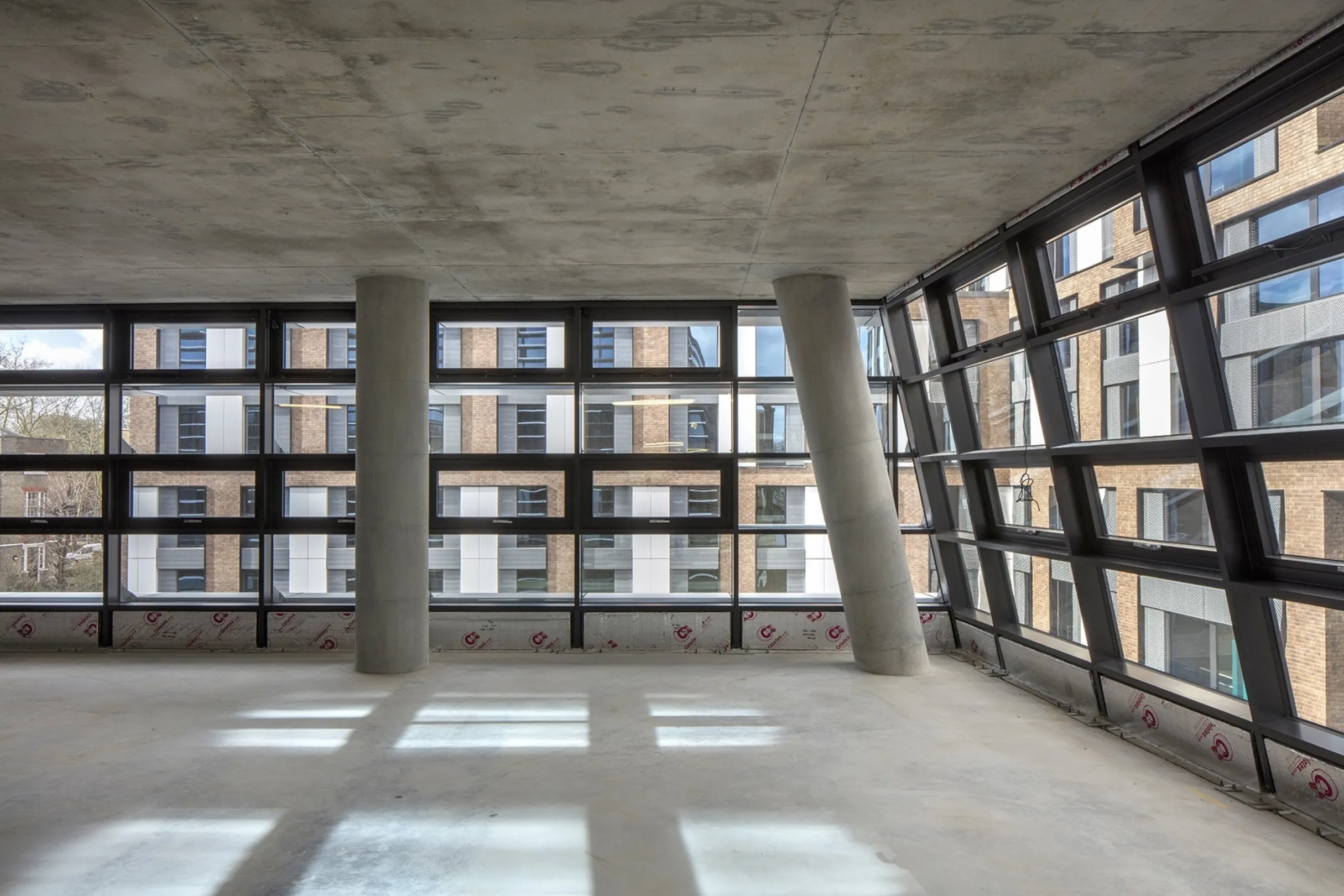 Interior detail showing concrete pillars and sloping glazed walls. A building from the same development is visible outside and shadows are cast on the floor.