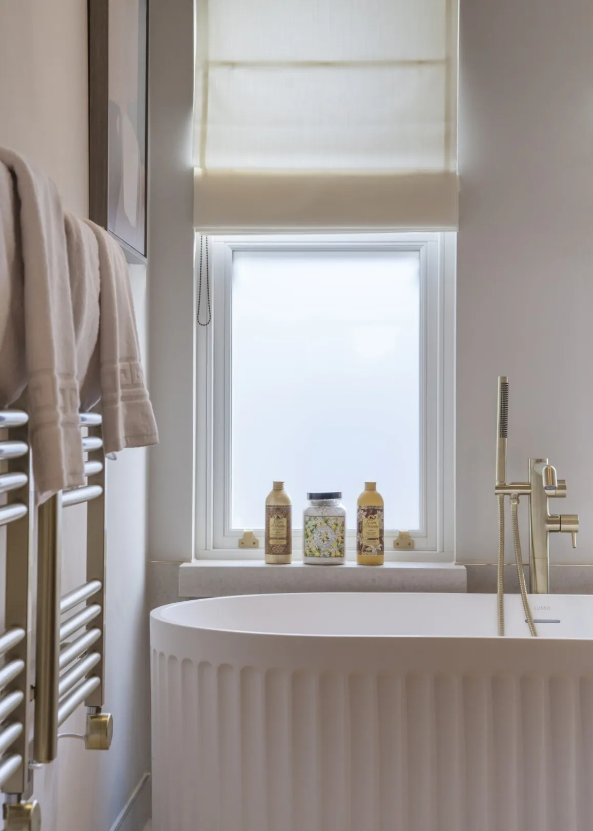 Master bathroom detail: A sculptural fluted freestanding bath with a floor-standing brass mixer sits beneath a softly lit sash window.