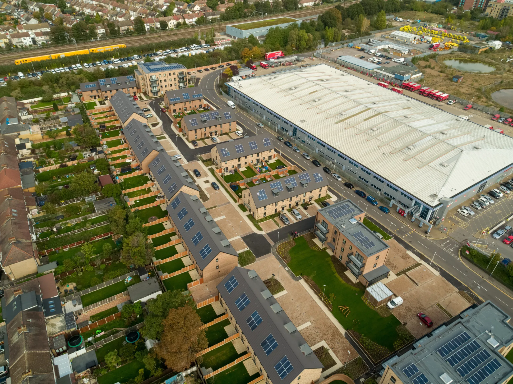 Arial view of multiple rows of modern terraced homes built in brick with dormer windows, parking in front and gardens to the rear