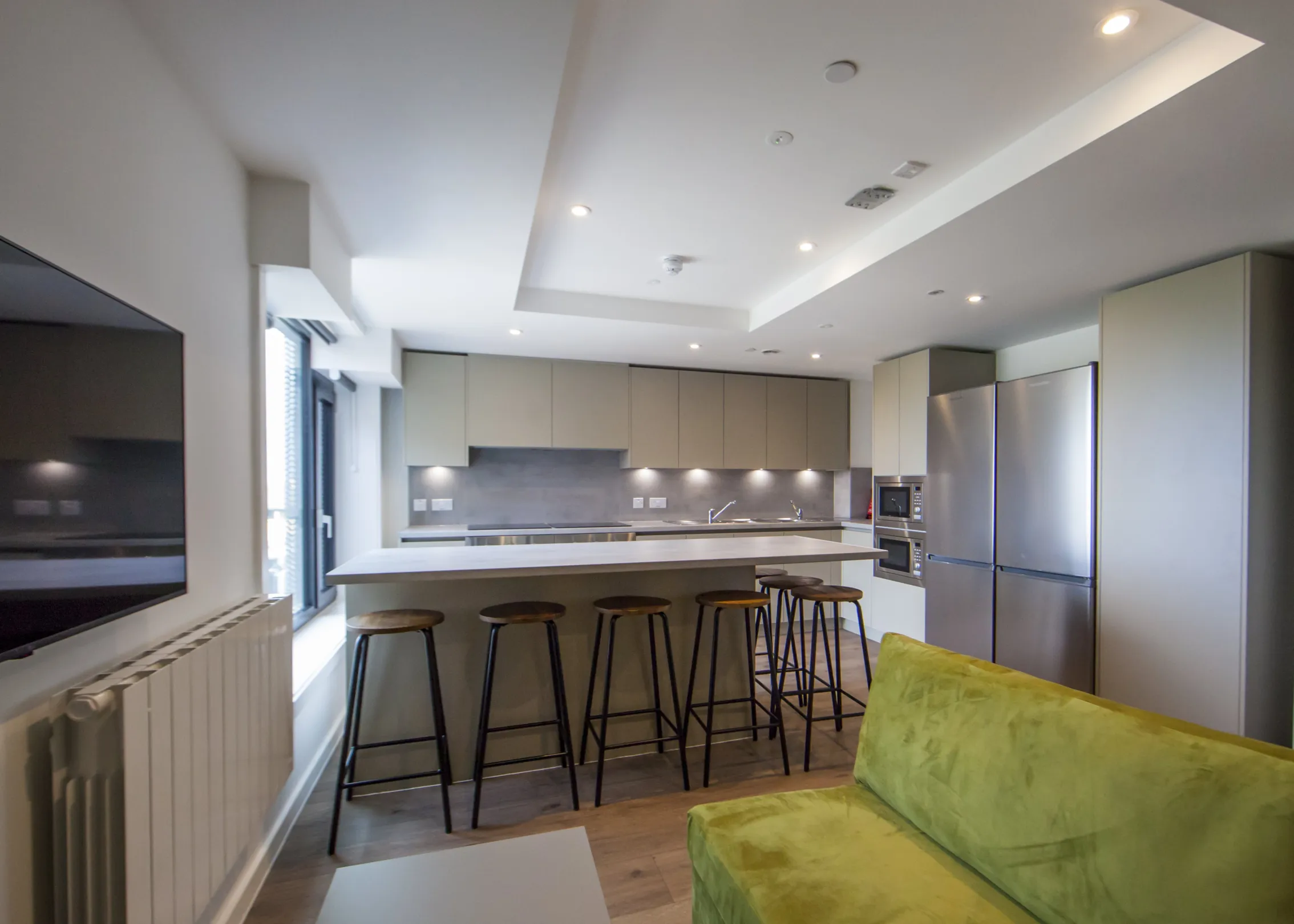 A shared kitchen area with eight stools around a breakfast bar, with sofa and tv in the foreground