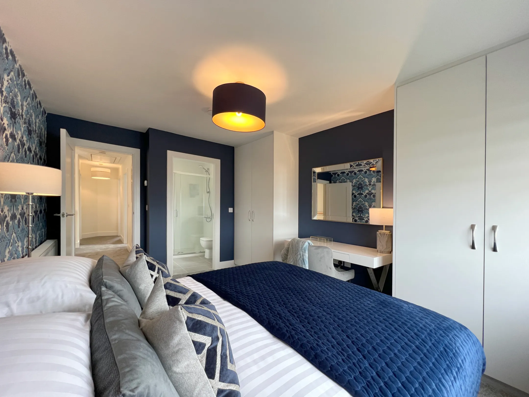 A modern double bedroom with navy wallpaper, built in white wardrobe and dressing table with mirror above