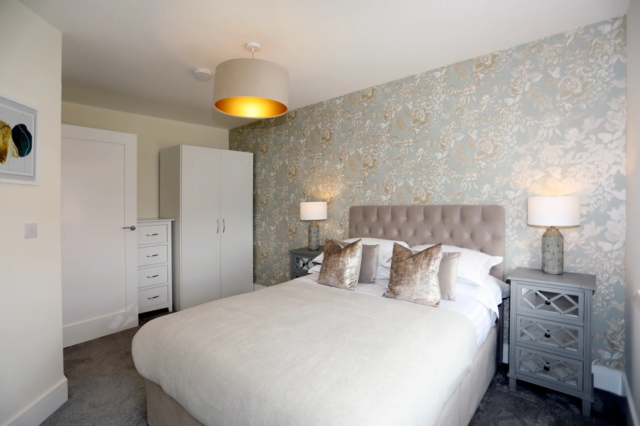 A modern double bedroom with textured wallpaper, freestanding white wardrobe and tallboy unit, dressing table with mirror above