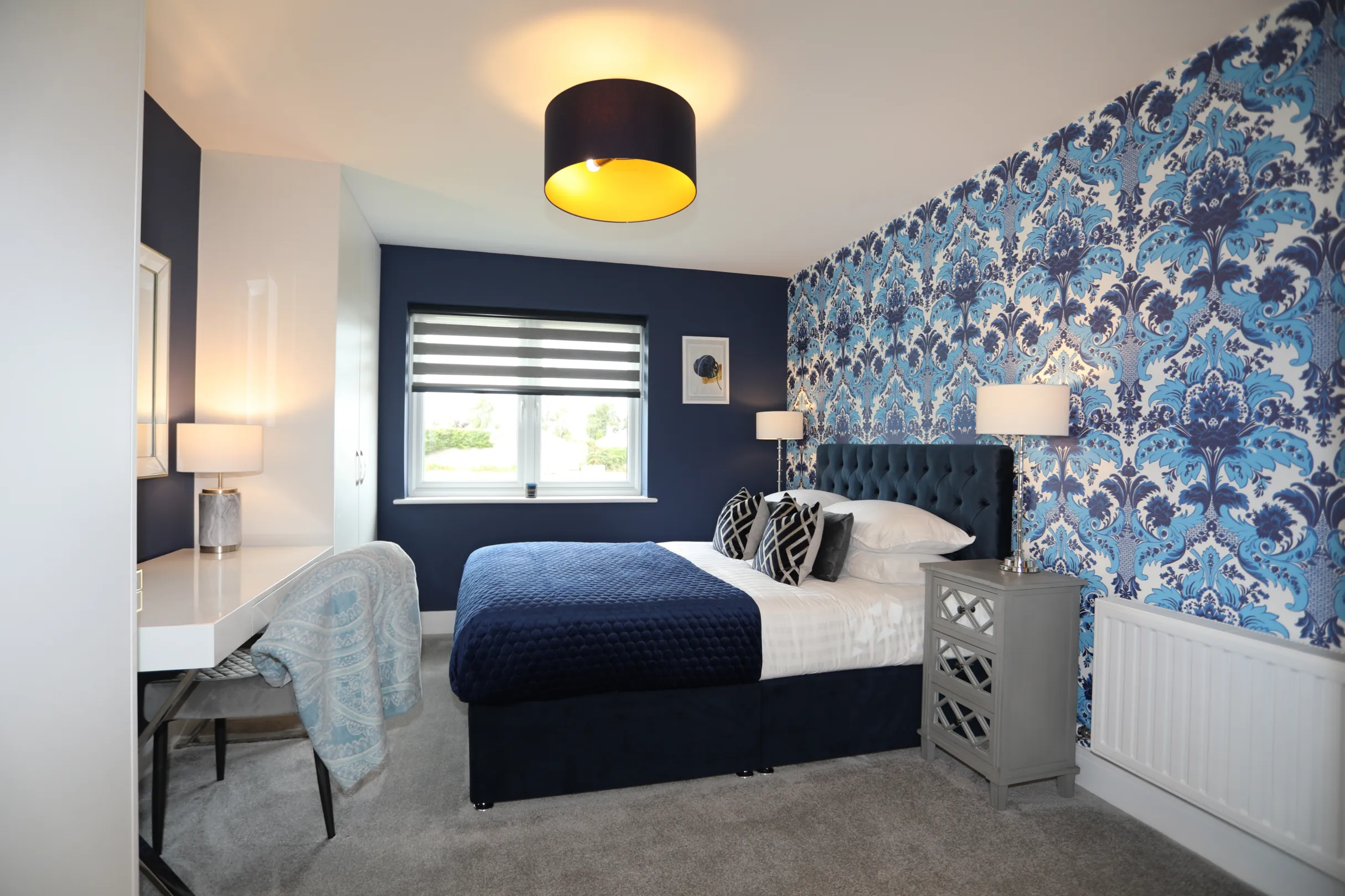 A modern double bedroom with navy wallpaper, built in white wardrobe and dressing table with mirror above