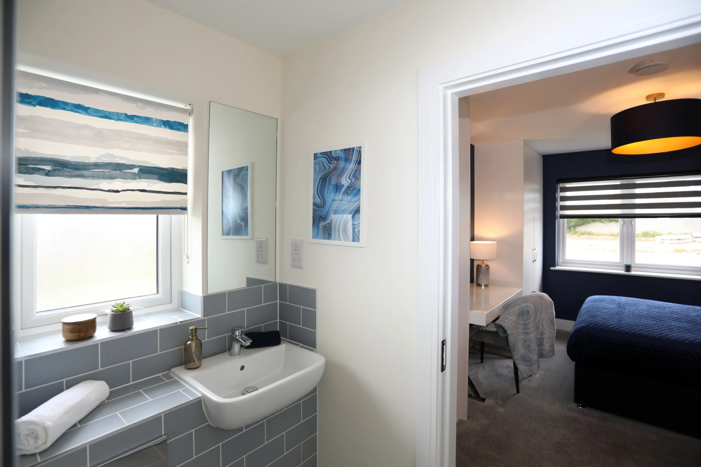 A master ensuite with light blue brick tiles, a white basin and WC, with a large mirror above the basin with a view back towards the bedroom