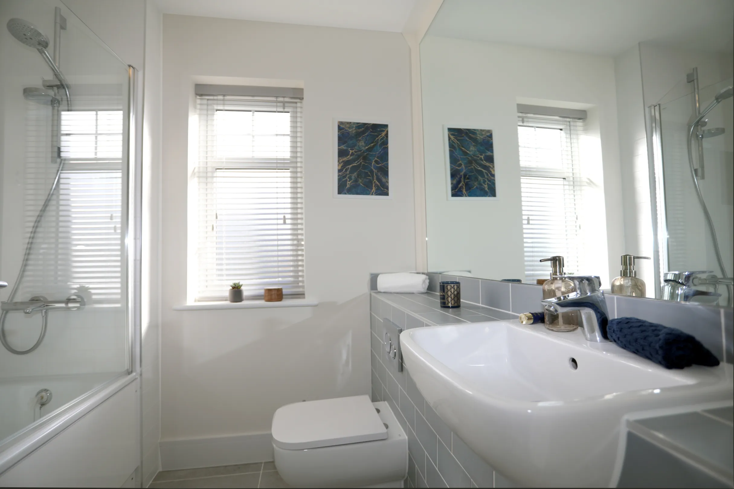 A main bathroom with light blue brick tiles, a white basin and WC, with a large mirror above the basin and a shower over bath