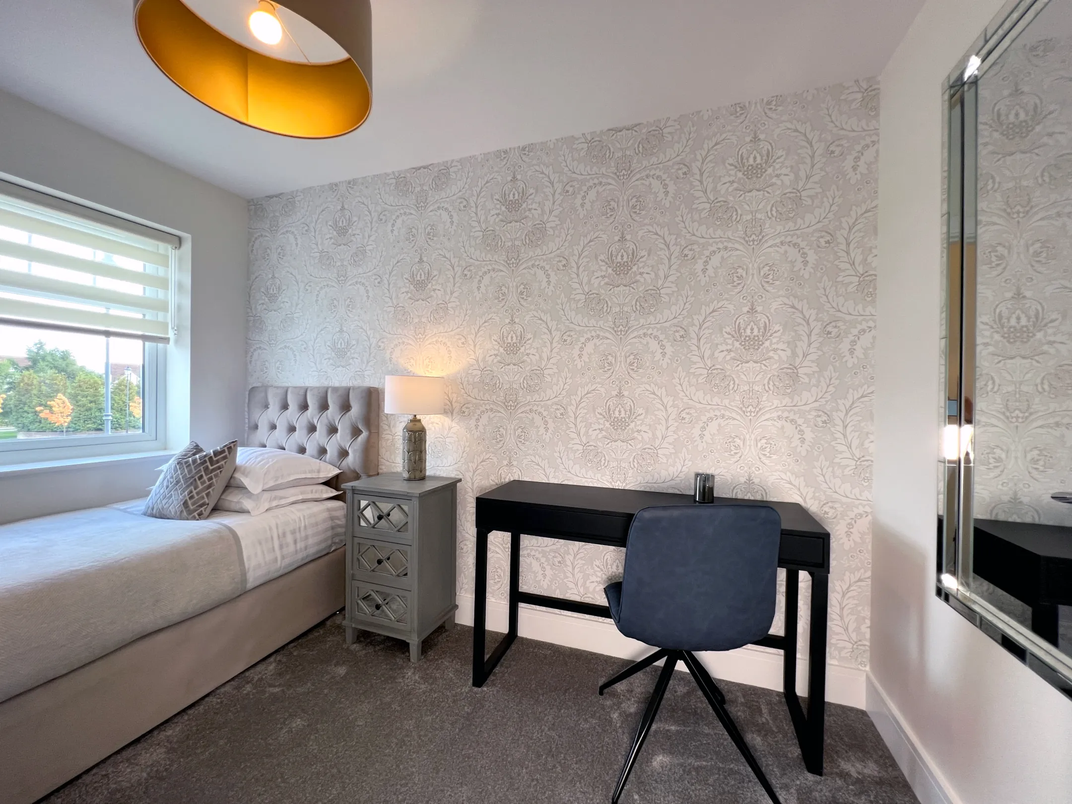 A single bedroom with taupe wallpaper on a feature wall, taupe bed through and dressing table with chair