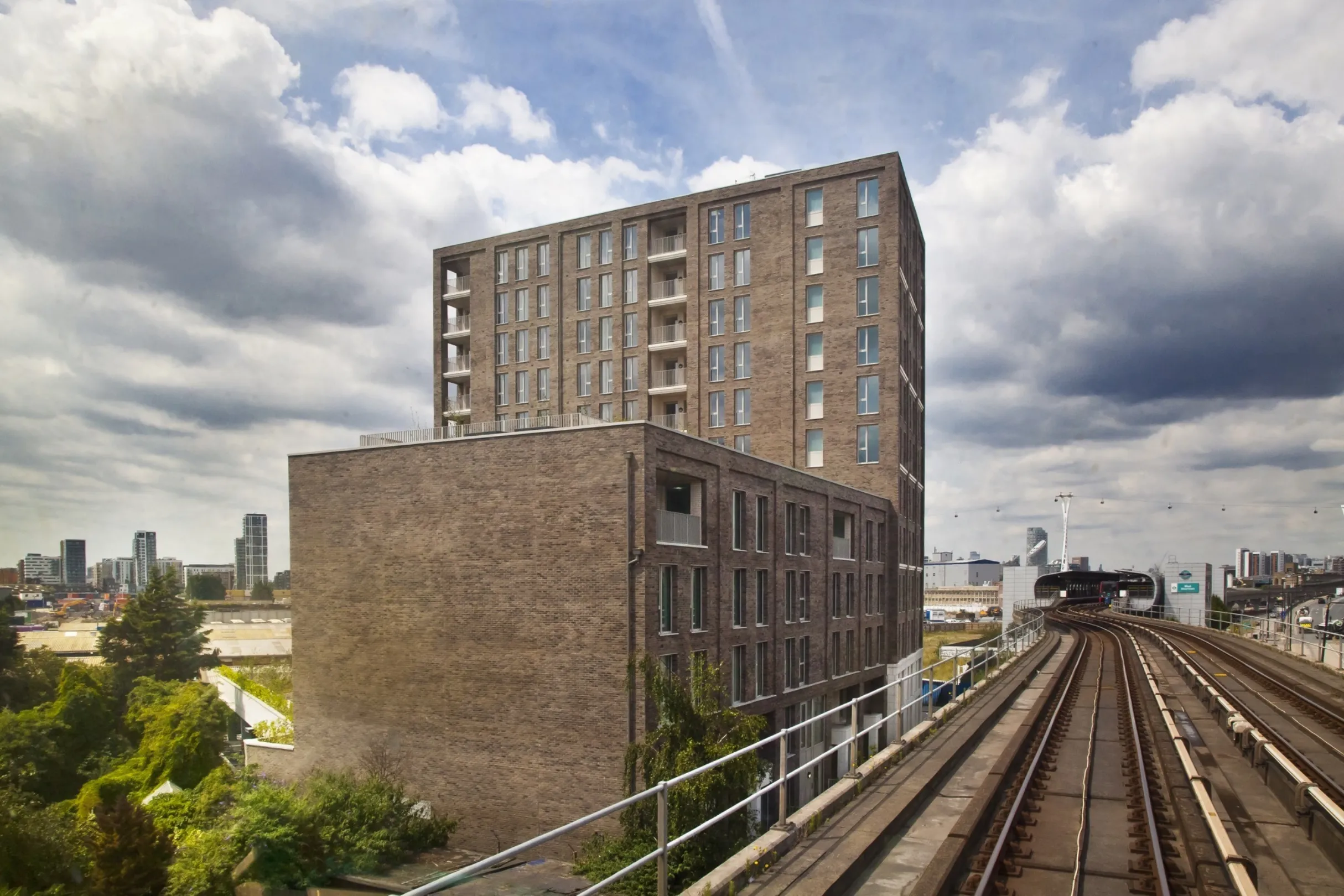 Detail modern multi storey residential building with balconies next to the Docklands Light Railway (DLR) track that leads into a station. The London cable car is in the far distance.