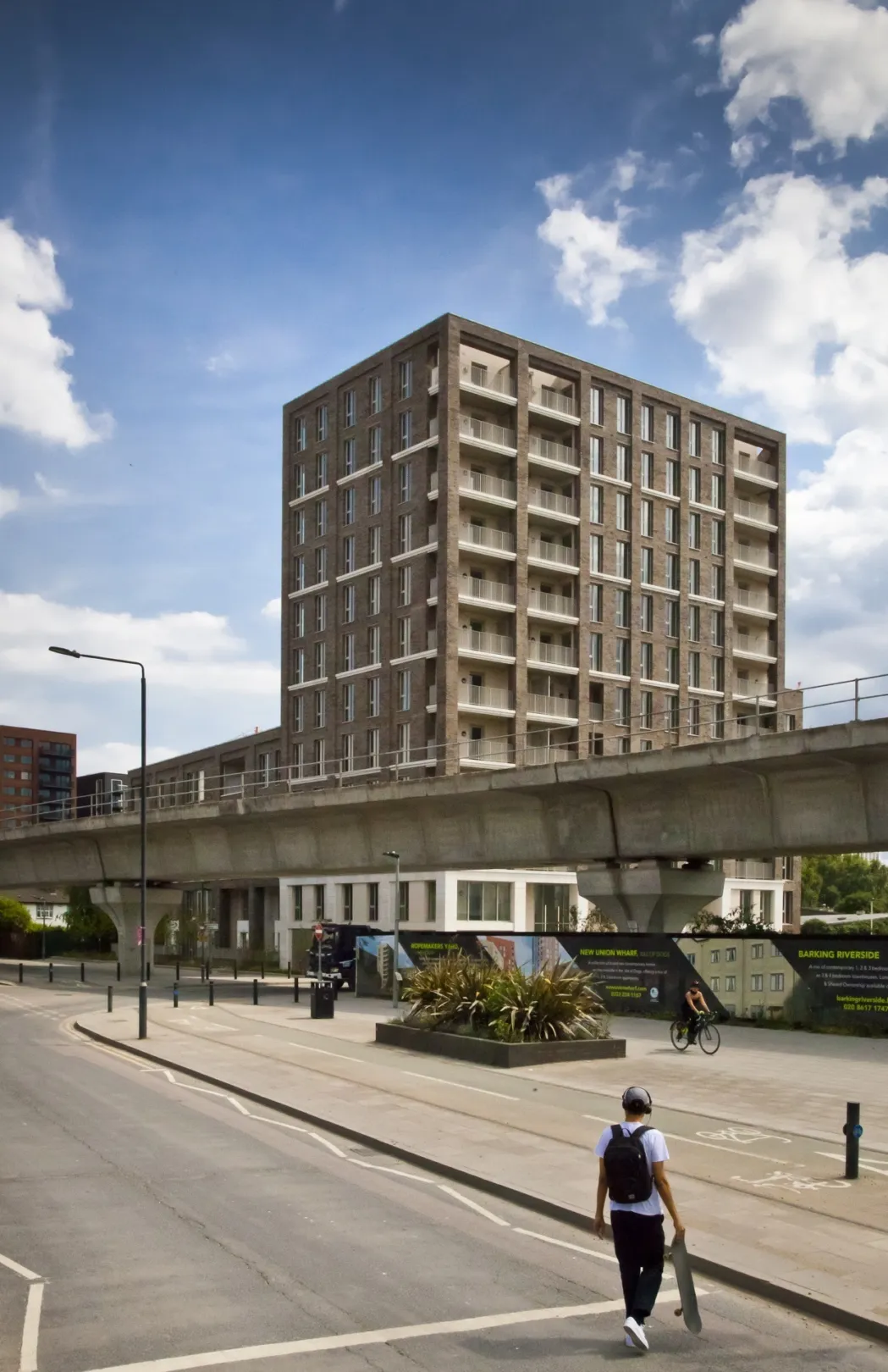 View from ground level towards a modern multi storey residential building with elevated train track, cycle path, wide pavement and street planting in the foreground.