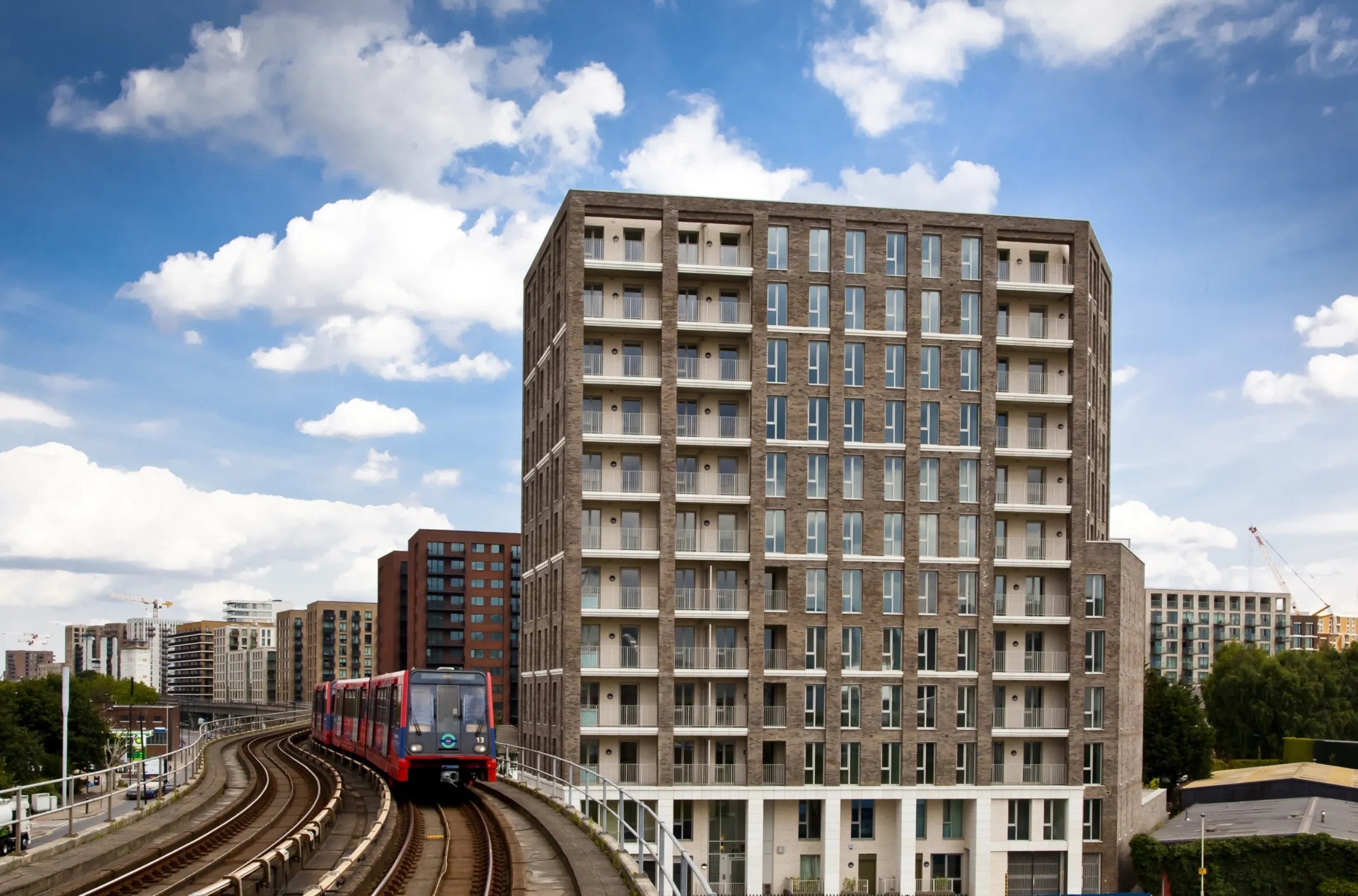 A modern multi storey residential building with balconies next to the London Docklands Light Railway (DLR) as a DLR train rolls along the track. Similar buildings stretch into the distance.