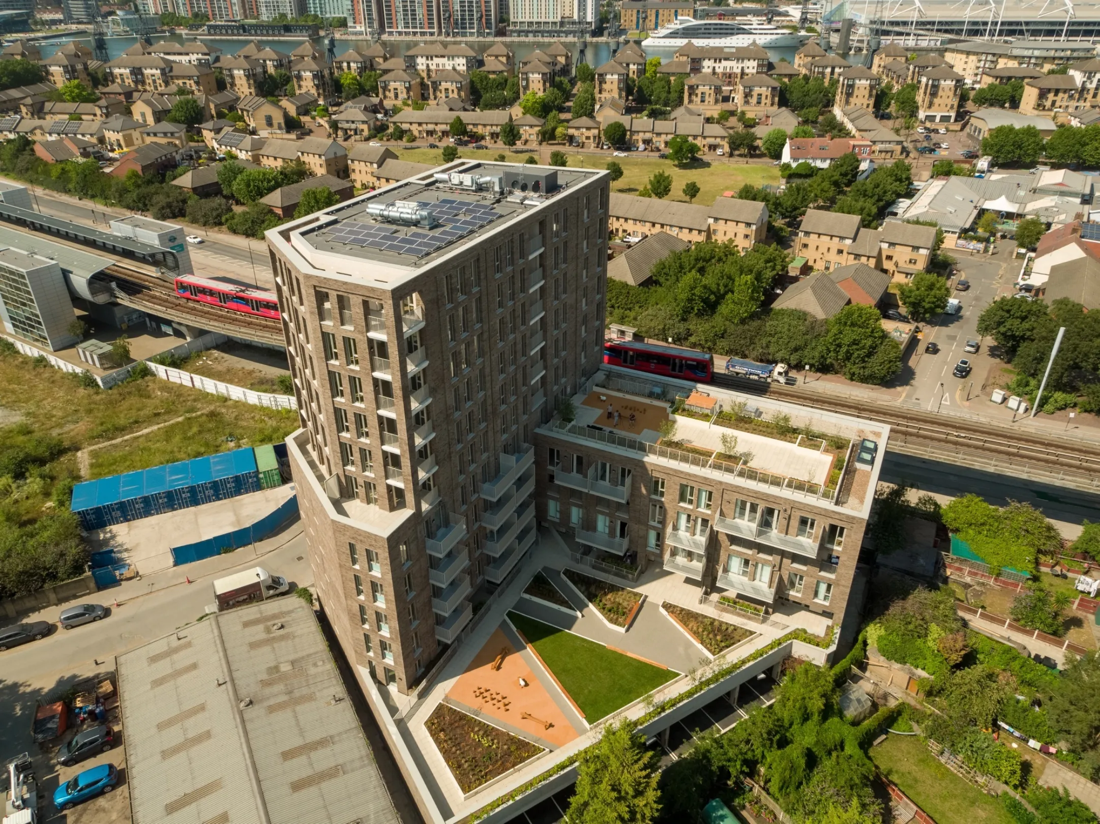 Arial view of a multi-level, multi story residential development showing solar panels on the highest roof and two levels of communal roof gardens. The docklands light railway (DLR) is in the background along with smaller homes and  luxury yacht moored in a basin in the distance.