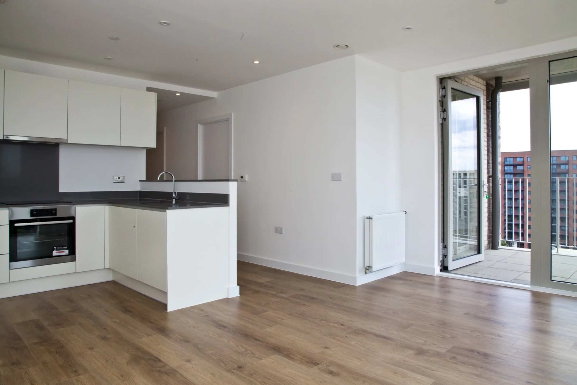 An empty modern kitchen and living area with a glazed wall that leads to a balcony overlooking more residential buildings.