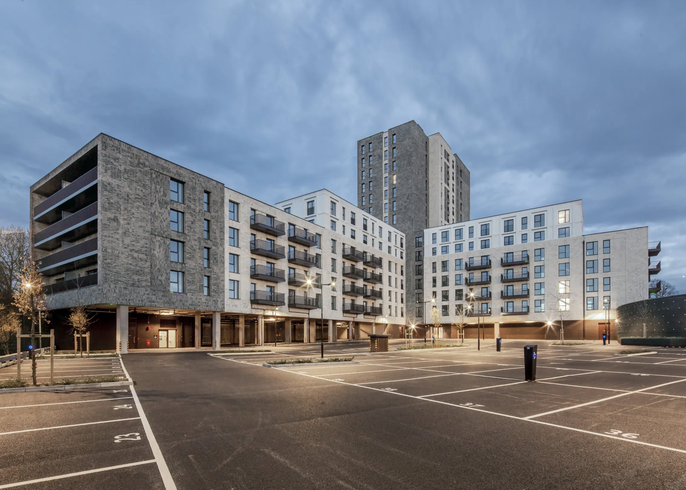 A modern residential building with different numbers of storeys in different parts. An illuminated parking area is show in the foreground.