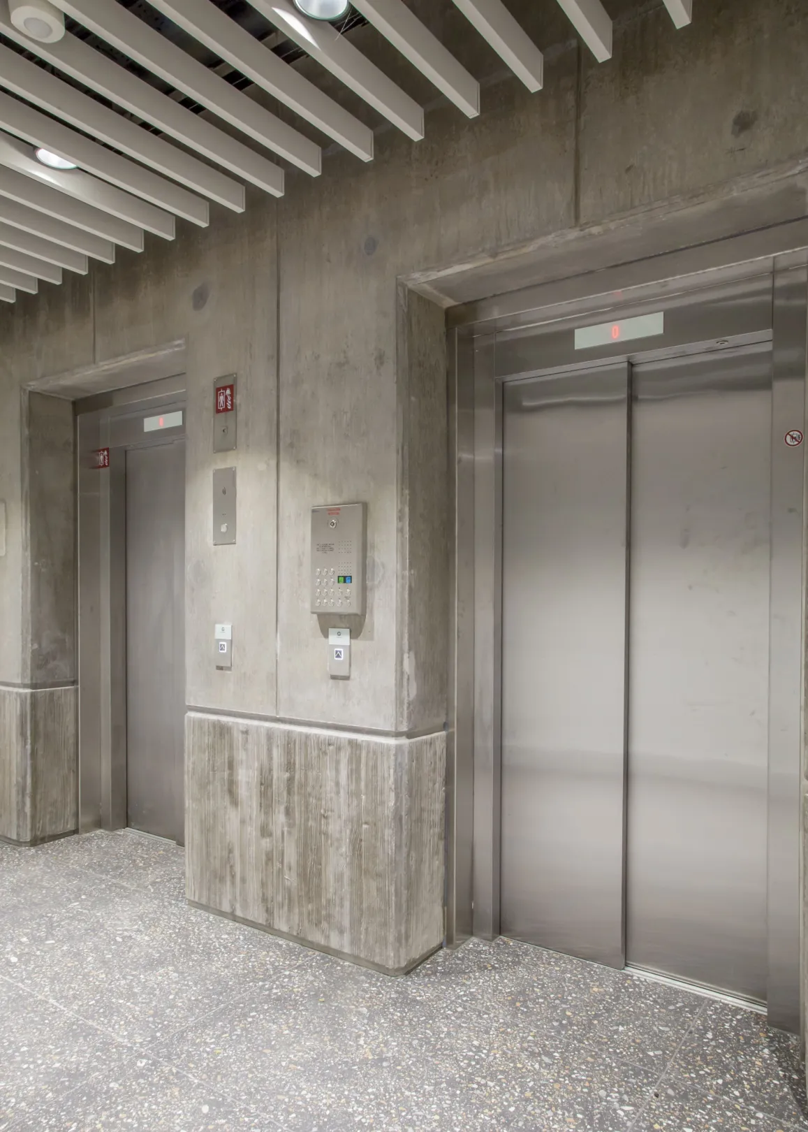 Entrance to two internal elevators with exposed concrete walls and suspended acoustic baffles on the ceiling.