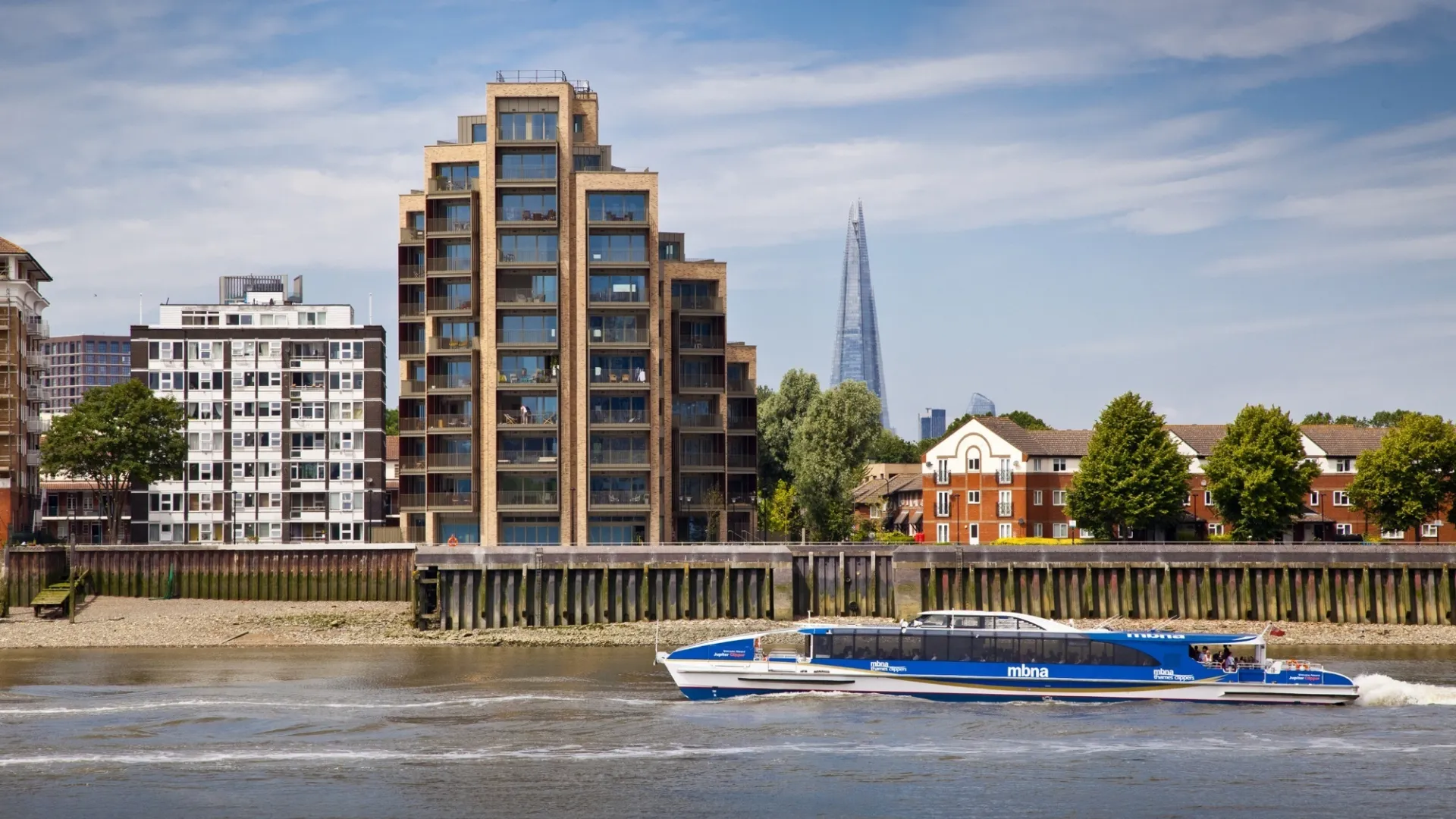 A modern multi storey residential building viewed from the River Thames as a passenger vessel speeds past. The Shard is in the far desistance.