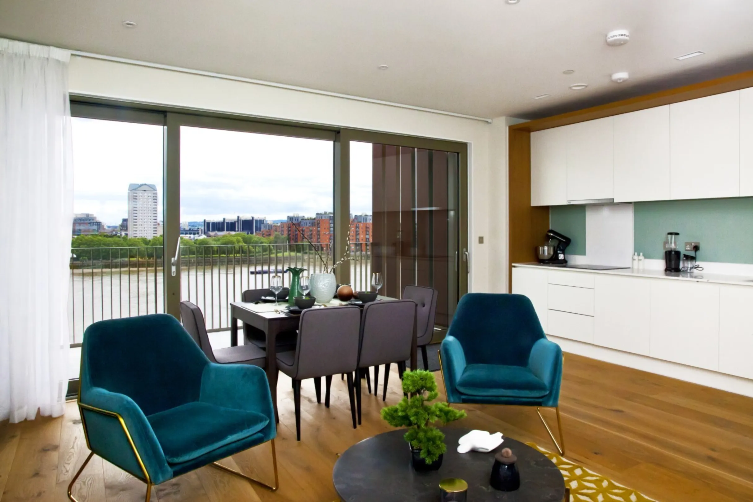 A white kitchen and wood flooring, with large balcony doors with a view of the river Thames looking towards Canary Wharf
