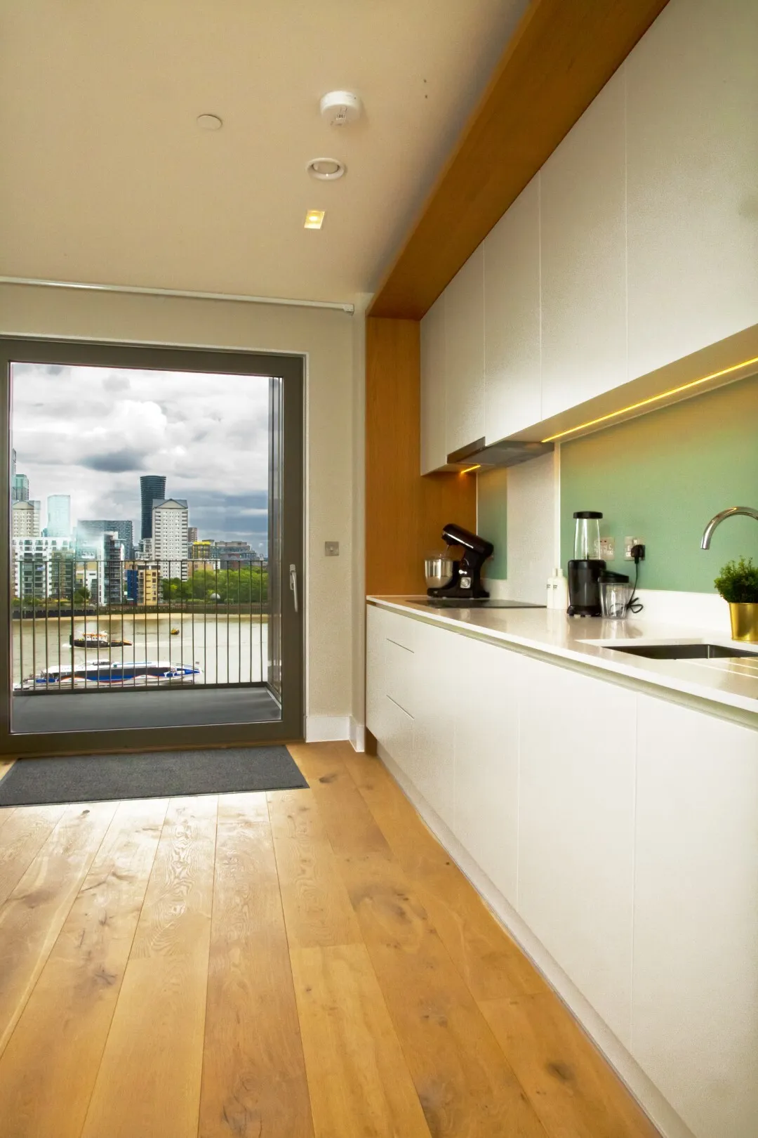 A white kitchen and wood flooring, with large balcony doors with a view of the river Thames looking towards Canary Wharf
