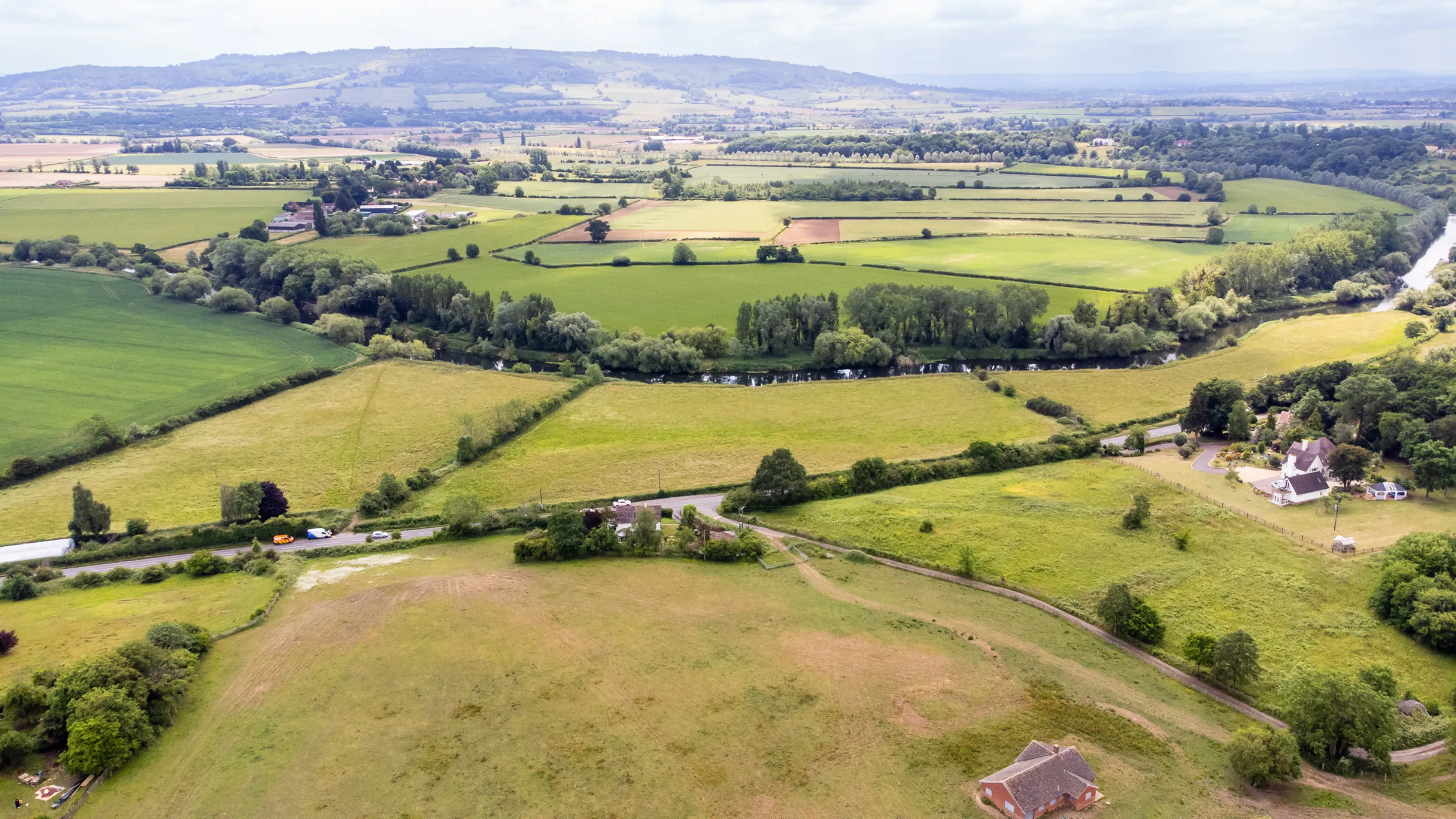 An aerial view captures a sprawling landscape of green fields, a winding river, scattered trees, and distant hills under a cloudy sky.
