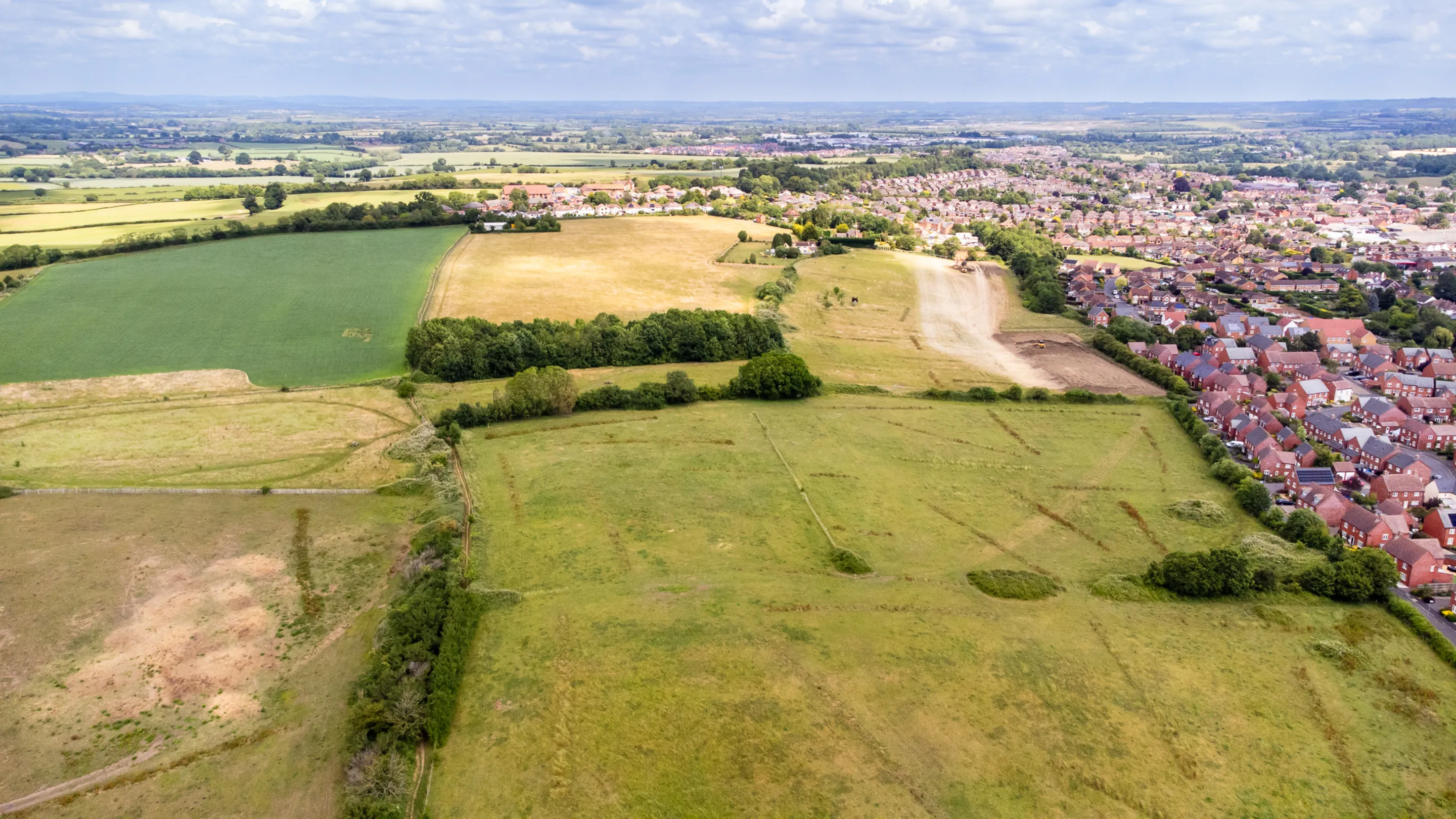 An aerial view reveals a patchwork of fields, ranging from lush green to golden brown, bordering a dense residential area under a cloudy sky.