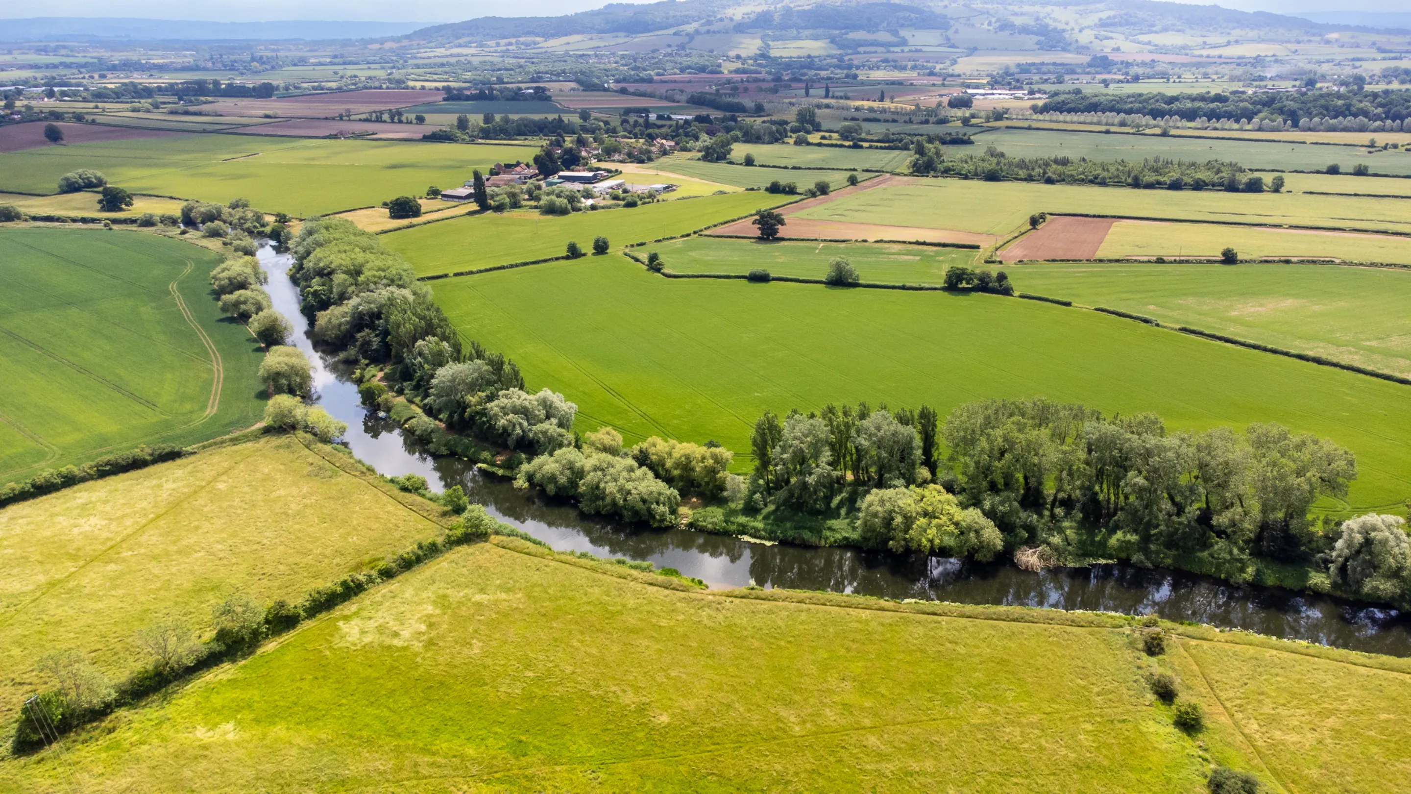 An aerial view shows a meandering river cutting through a landscape of vibrant green fields and scattered trees, with distant hills in the background.
