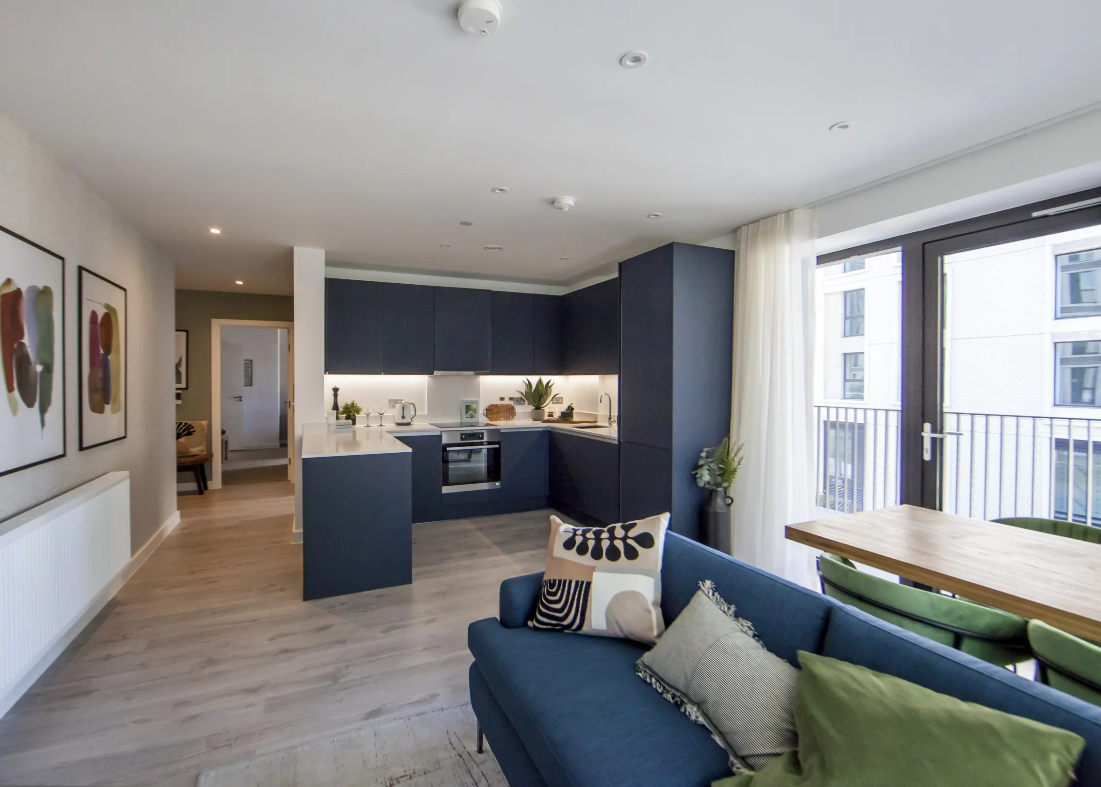 A modern apartment living space featuring a blue kitchen, blue sofa, and a dining area near a balcony.