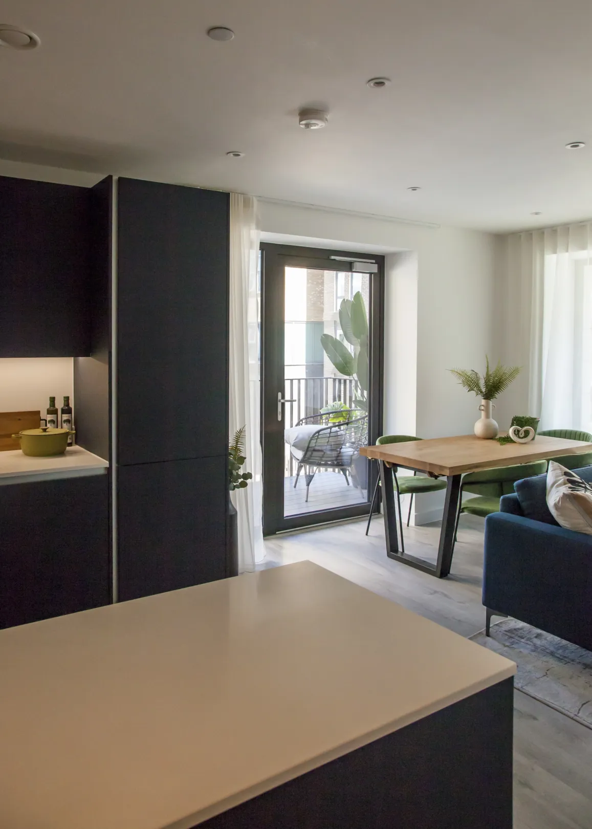 A modern apartment features a dark kitchen island, dining table, and a glimpse of a balcony with outdoor seating.