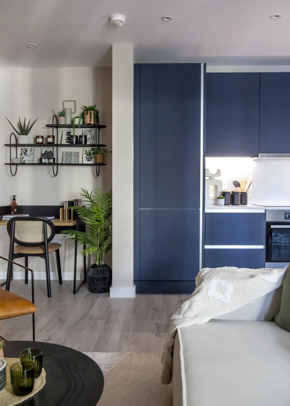 A compact and modern living space featuring a desk area with shelves, a sleek blue kitchen, and a cozy seating area with a round table.