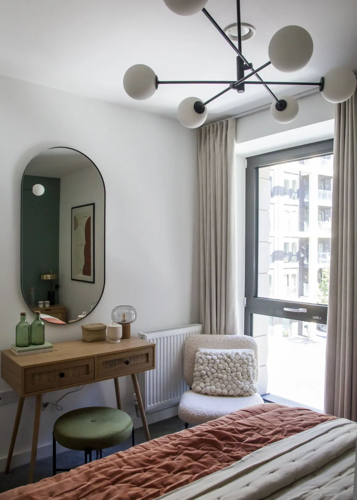A cozy bedroom with a vanity, oval mirror, a modern chandelier, and a plush chair next to a window.