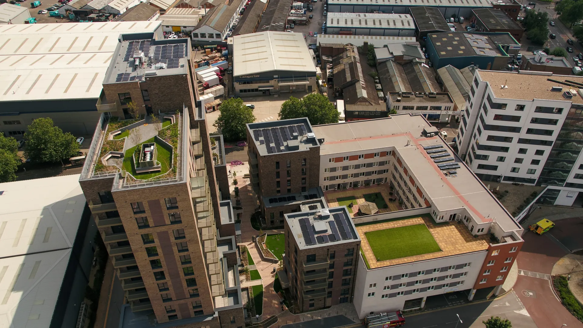 Arial view of a large residential development with multiple levels, rooftop garden and parking, sitting amongst other buildings of varying sized, ages and types