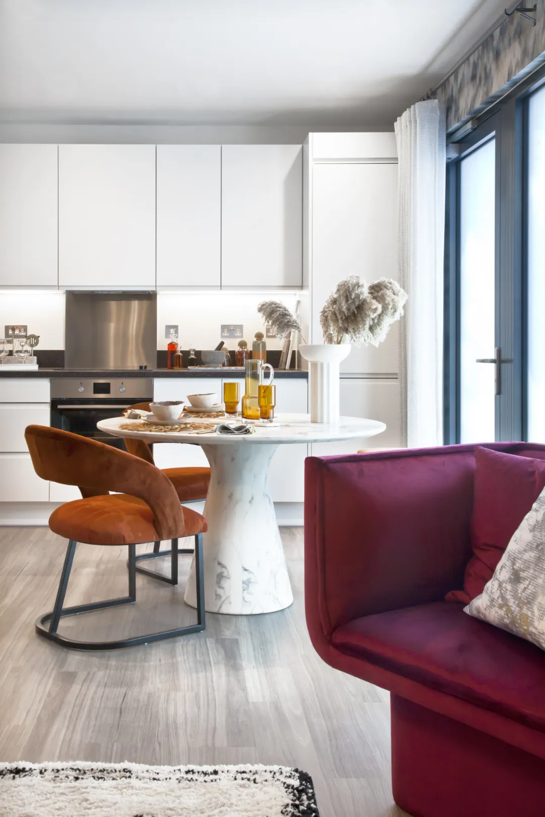 A modern kitchen dining room, with a white kitchen, integrated appliances and dark worktop.  A round dining table with organe chairs.