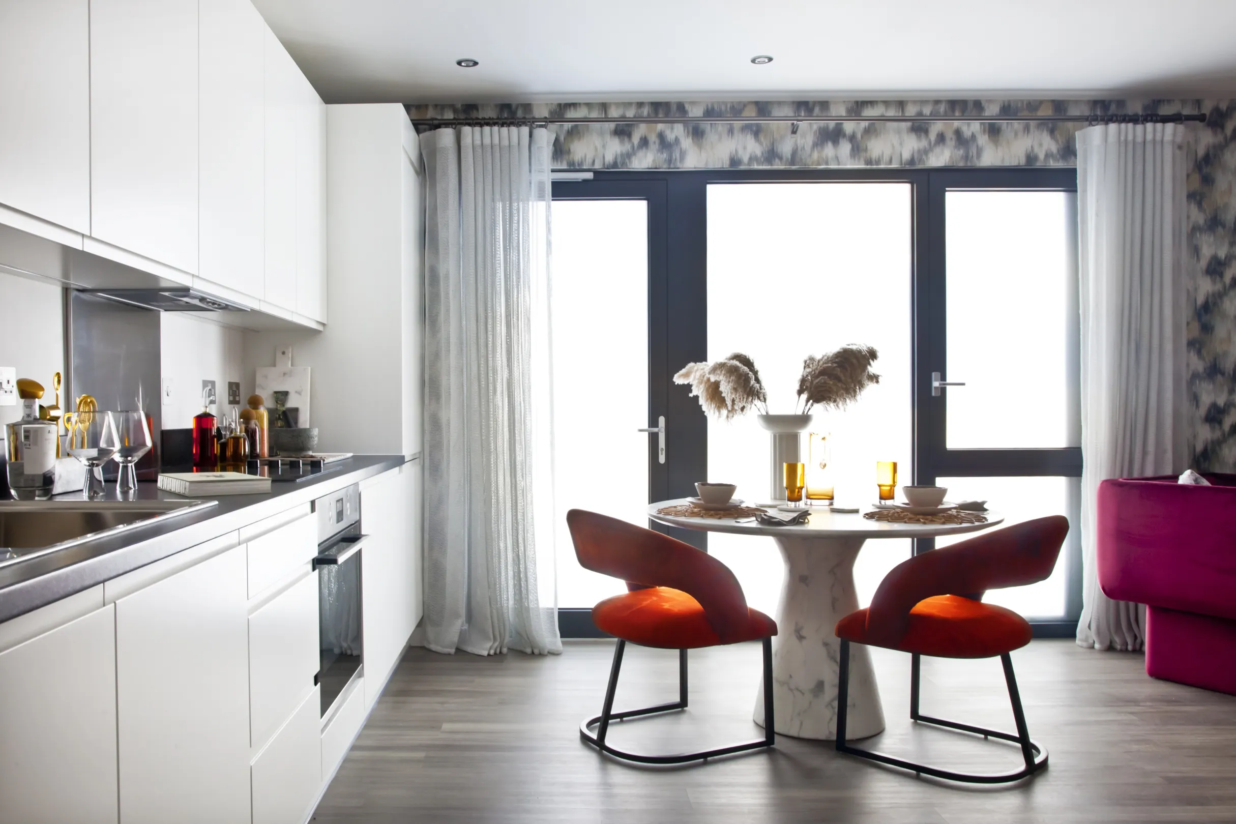 A modern kitchen dining room, with a white kitchen, integrated appliances and dark worktop.  A round dining table with organe chairs.