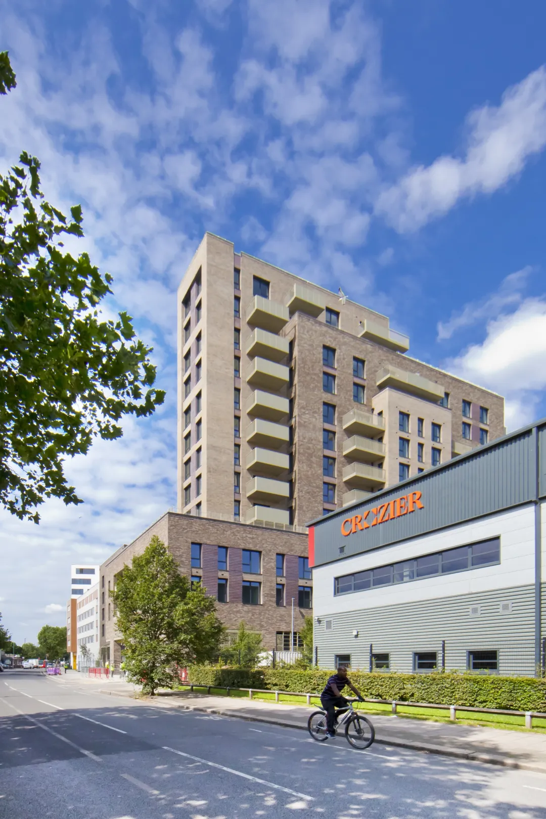 A man cycles past a modern multi storey building in summer with a smaller commercial building in the foreground
