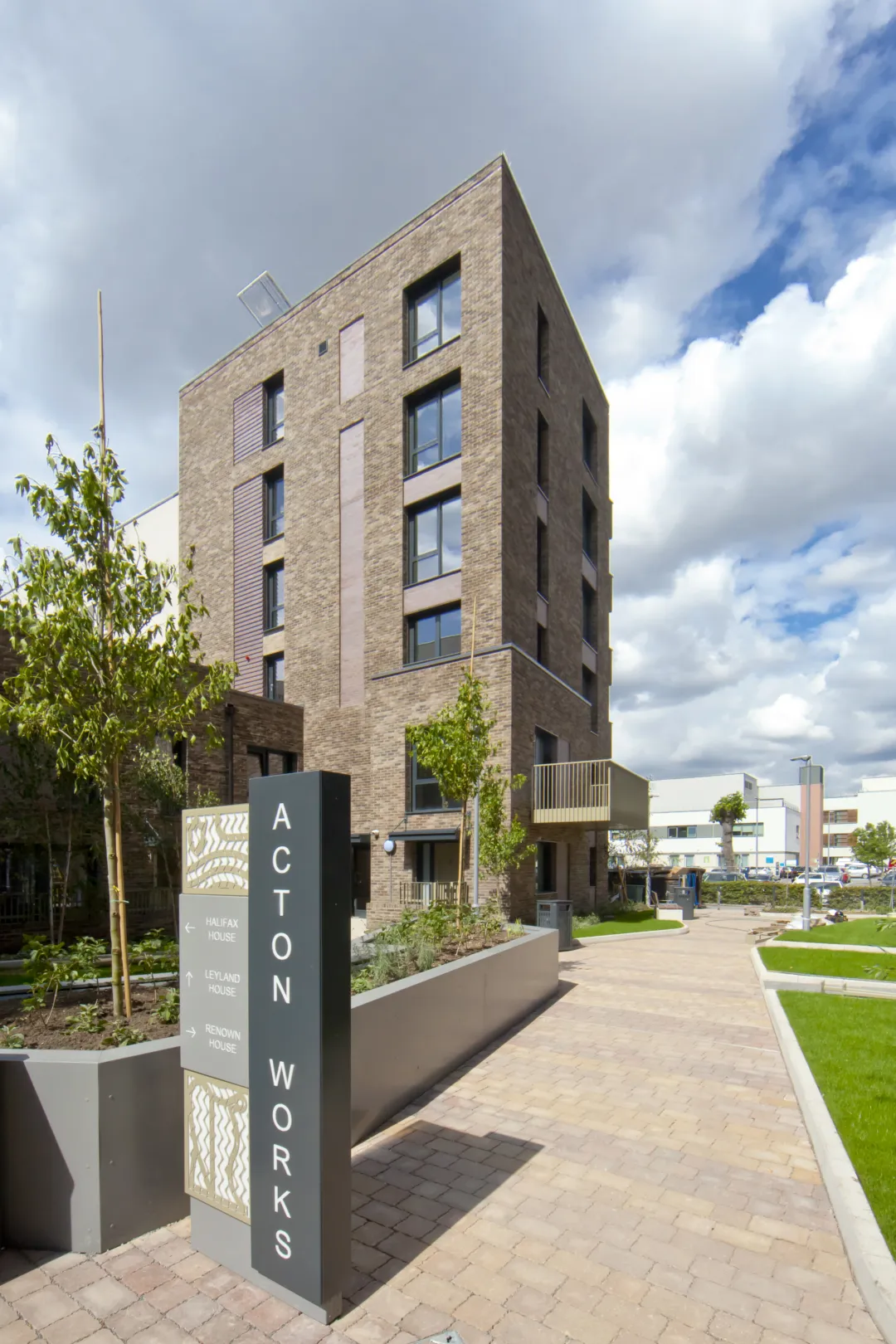 A modern brick facade residential building with lawned and paved grounds, and a wayfinding sign reading "Acton Works"