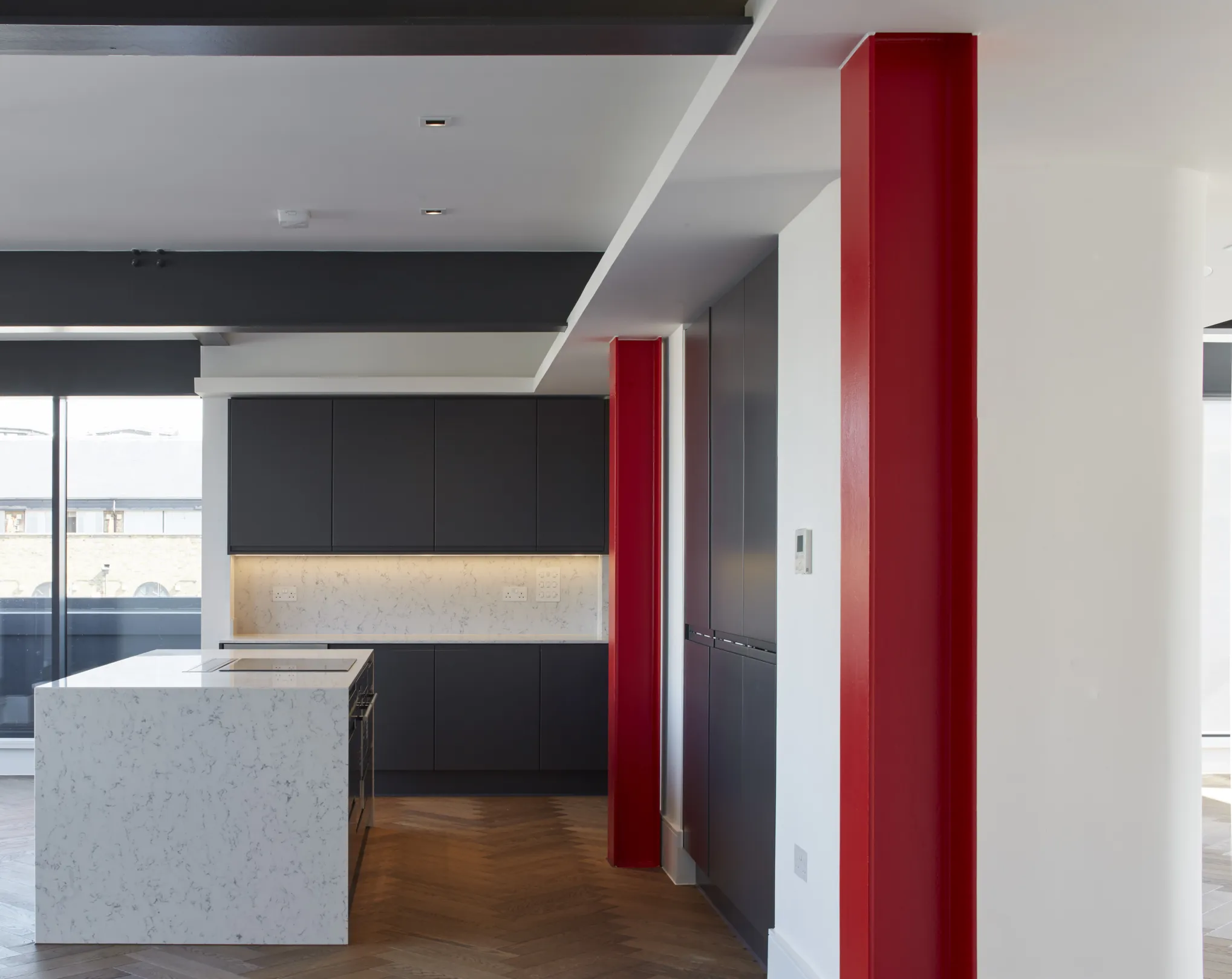 Modern large aparment, with grey kitchen, marble white worktop, integrated appliances, herrinbone flooring and red steelwork