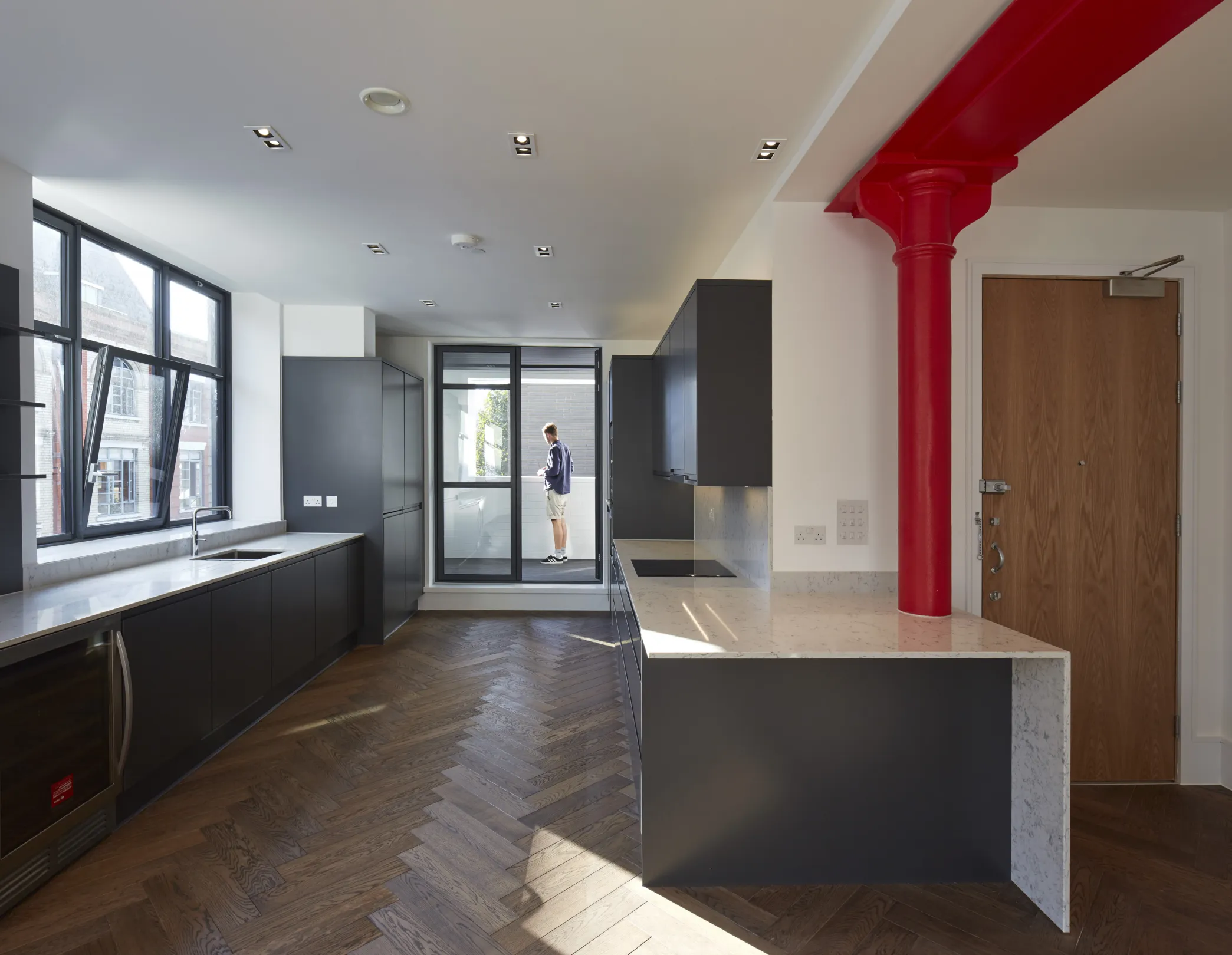Modern large aparment, with grey kitchen, marble white worktop, integrated appliances, herrinbone flooring and red steelwork
