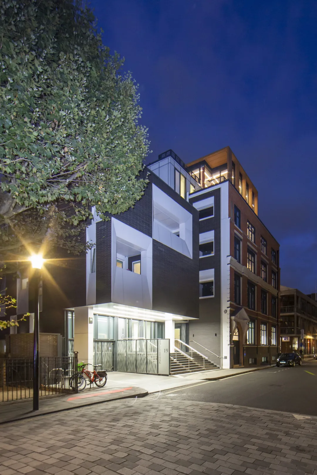 Modern office building entrance illuminated at night, abutting a residential building that forms part of the same development. Trees are in the foreground.