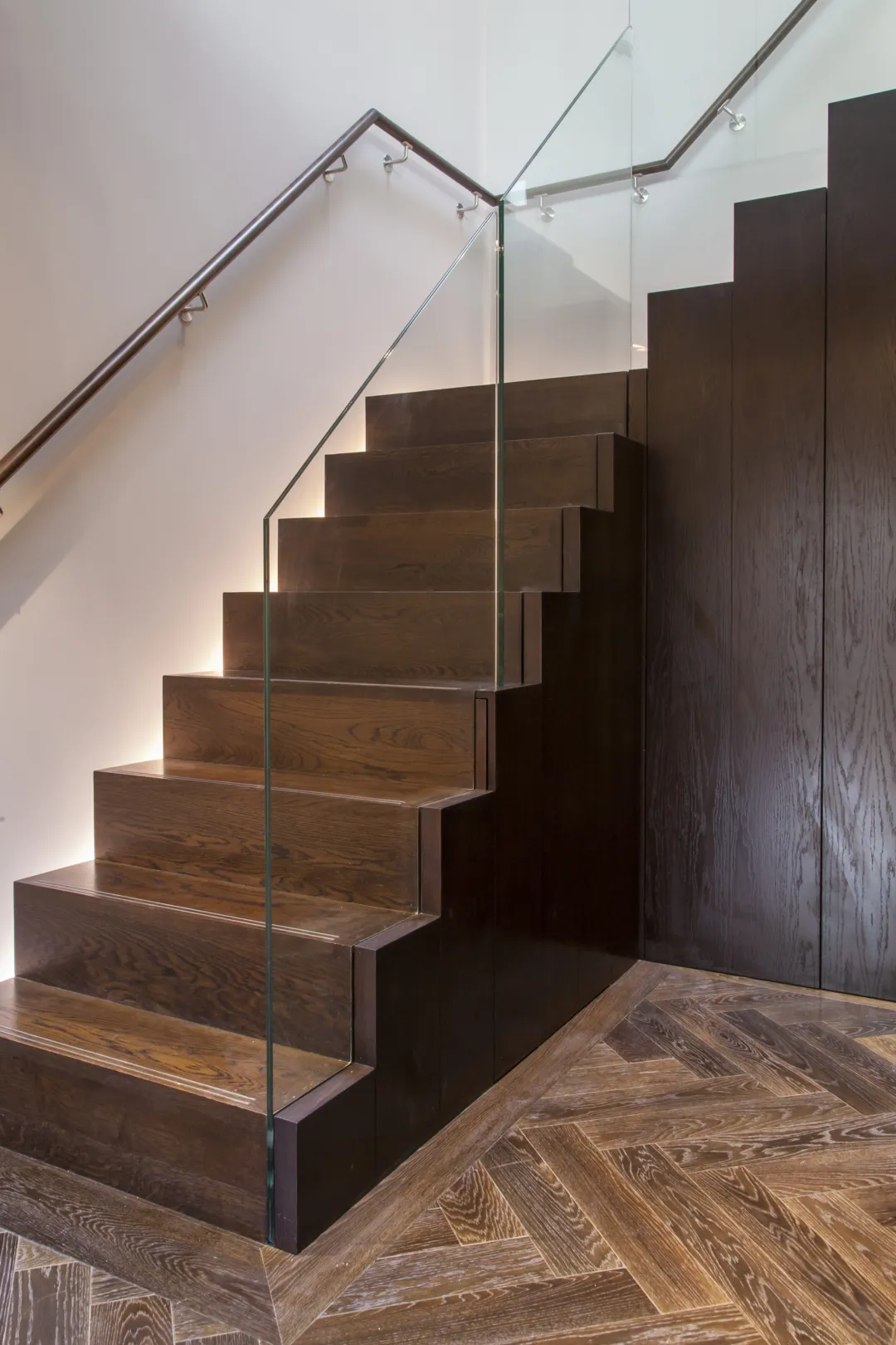 Timber clad staircase with glass balustrade and concealed lighting on the wall side and dark parquet floor