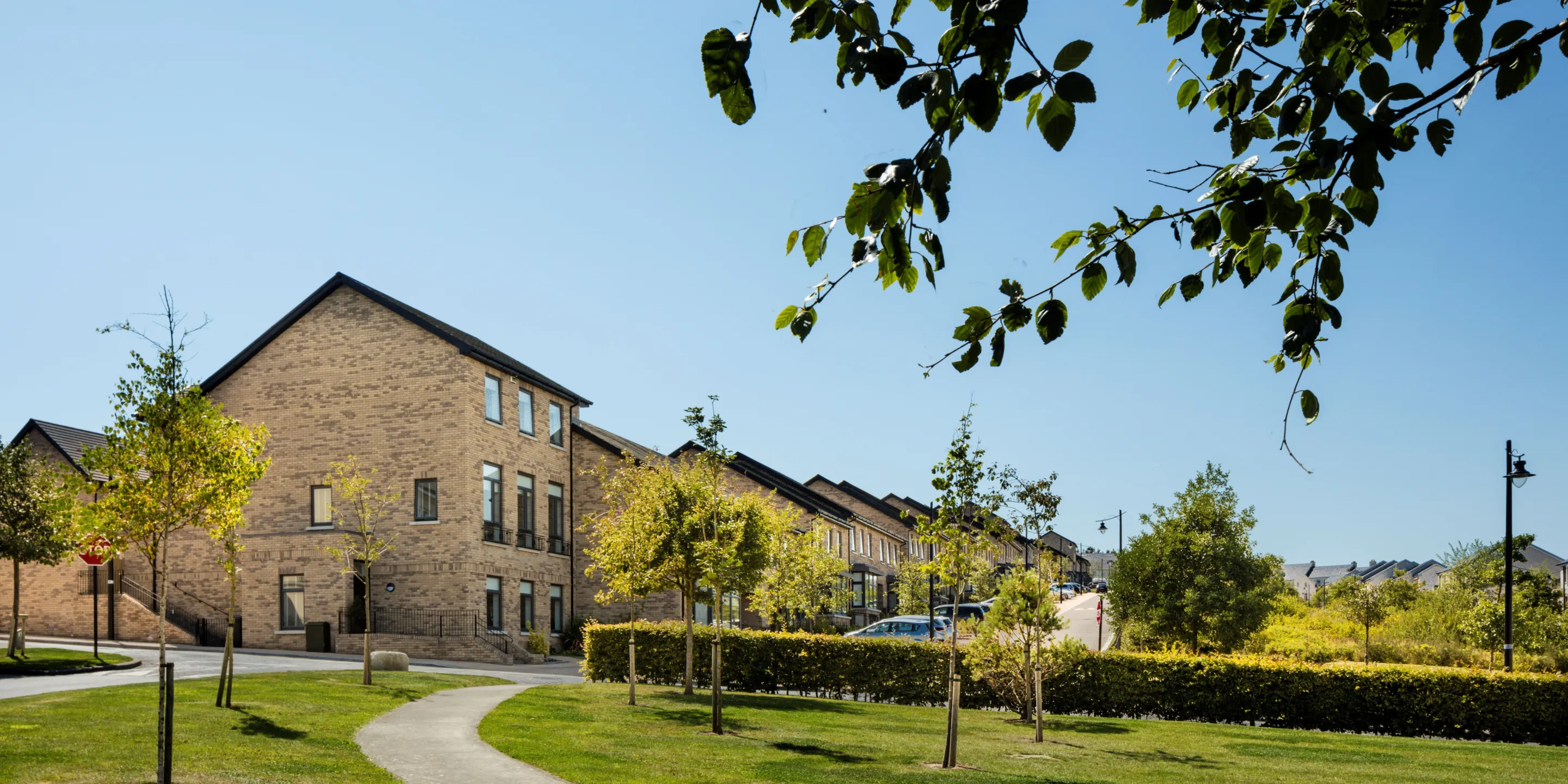 A row of terraced yellow brick houses with a large area of landscaping and footpath with trees