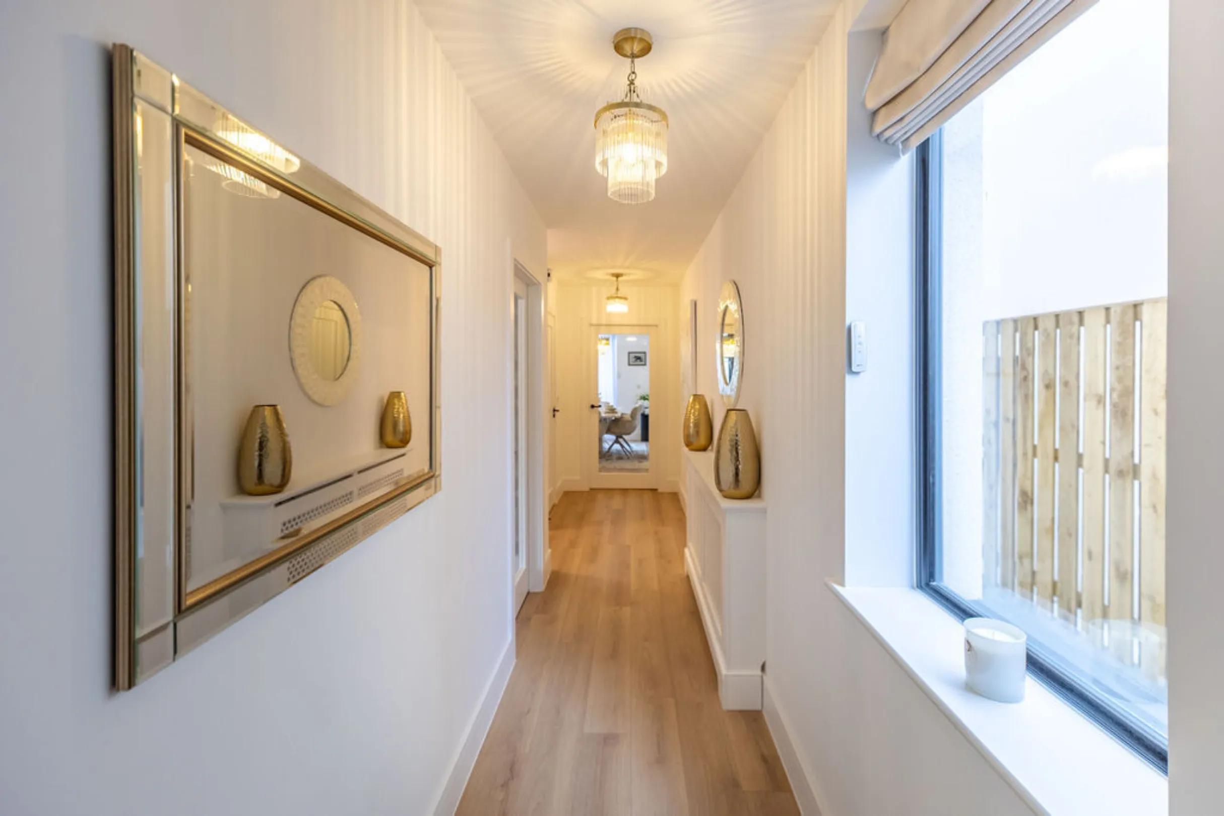 A well-lit, narrow hallway features light wooden flooring, white walls adorned with gold-accented mirrors and vases, and a large window