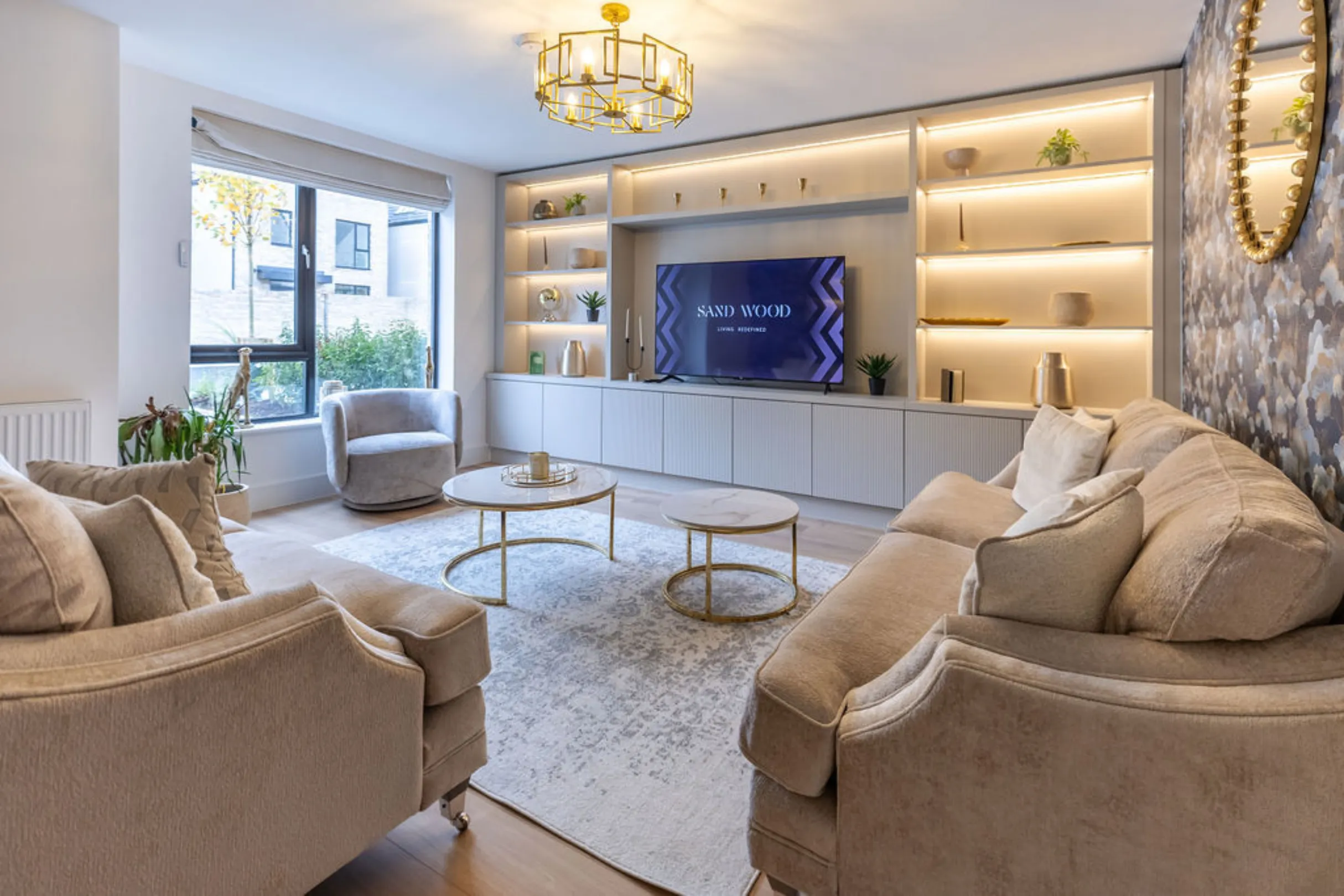 A stylish and inviting living room features comfortable, neutral-toned seating arranged around two round coffee tables on a patterned rug, complemented by an illuminated built-in media console and shelving, and ample natural light from a large window.
