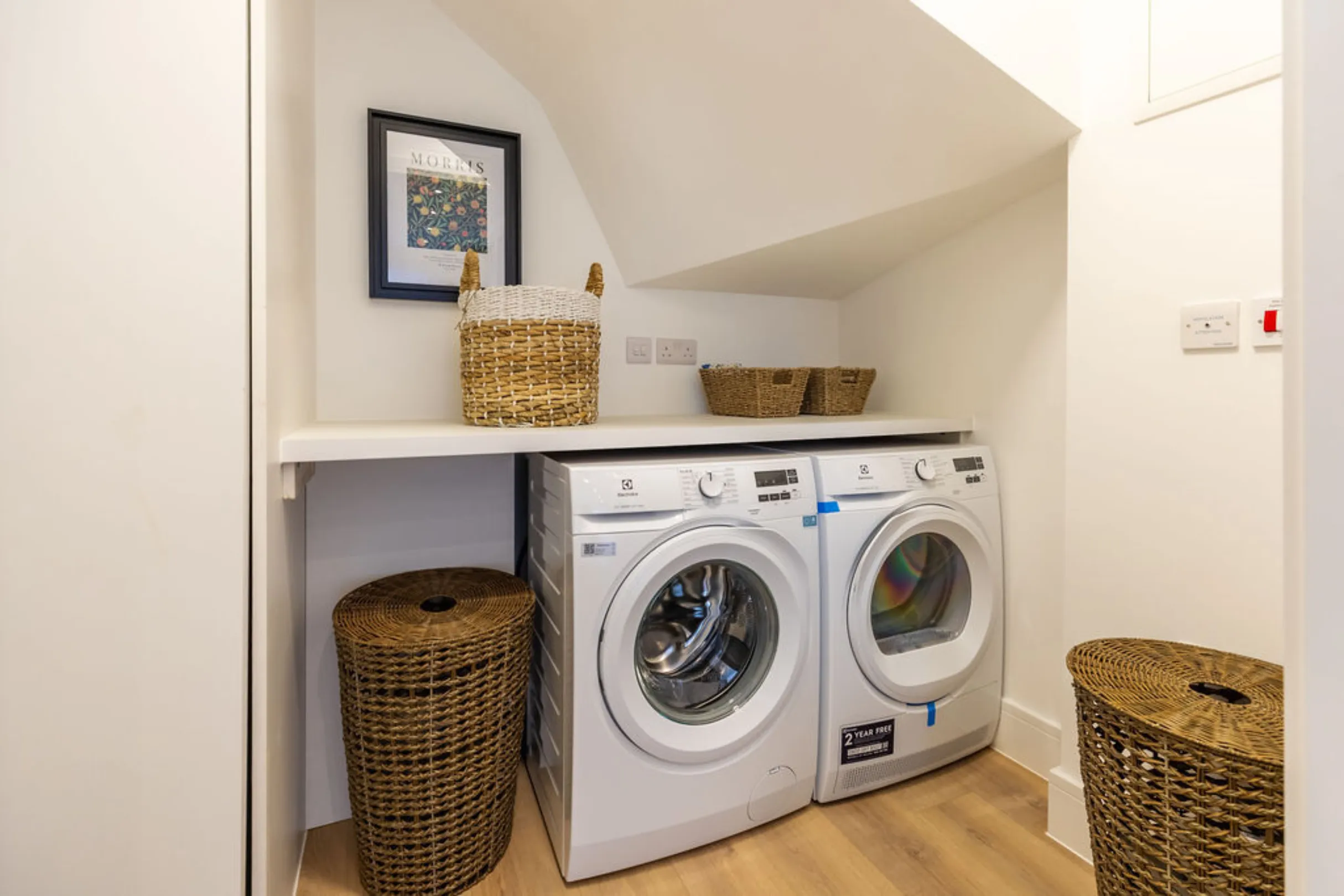 The image shows a laundry room with a washing machine and a dryer. There are also decorative baskets and a framed picture.